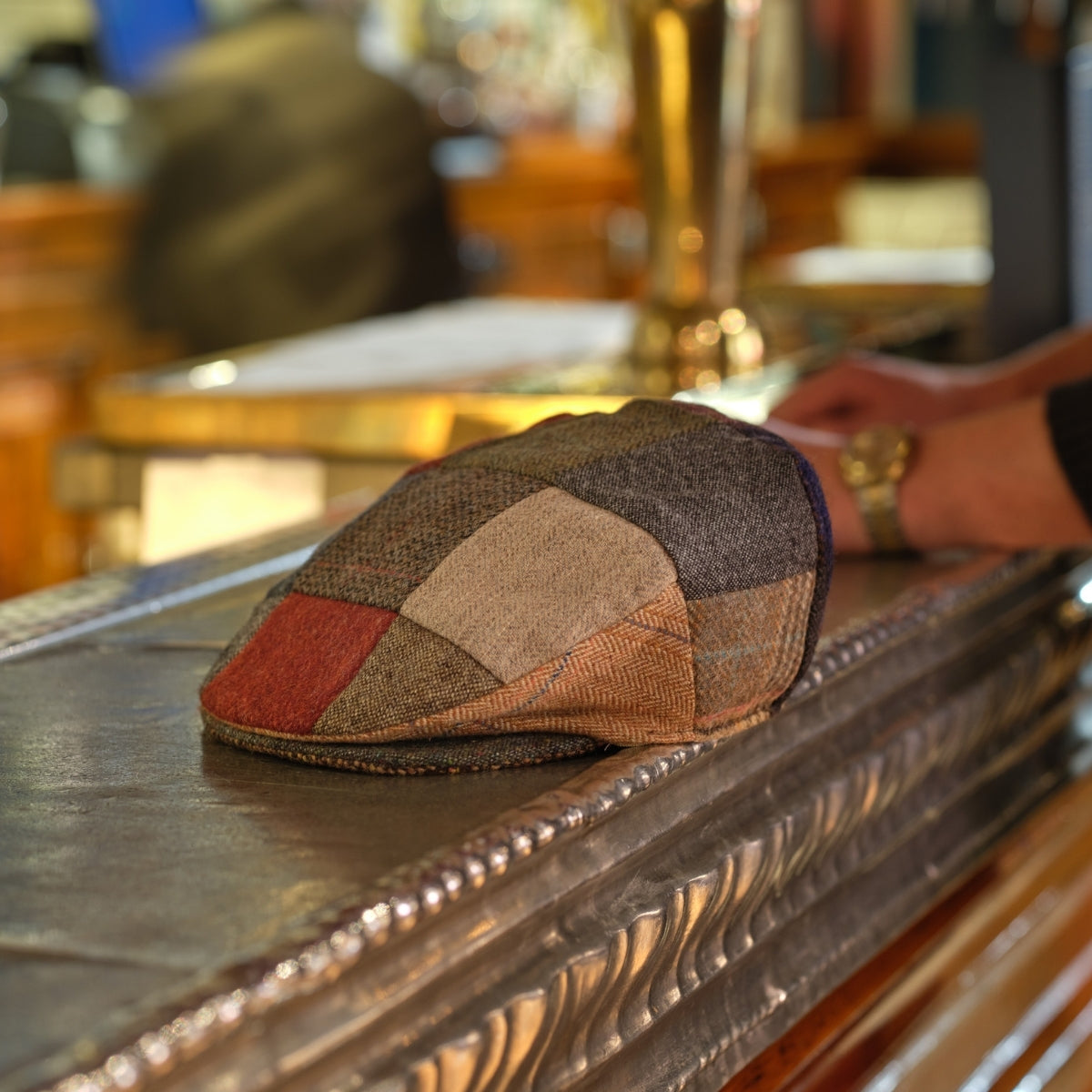 A patchwork flat cap rests on a polished metallic bar counter. In the background, a person’s arm and hand are visible, along with blurred, warm-toned bar decor.