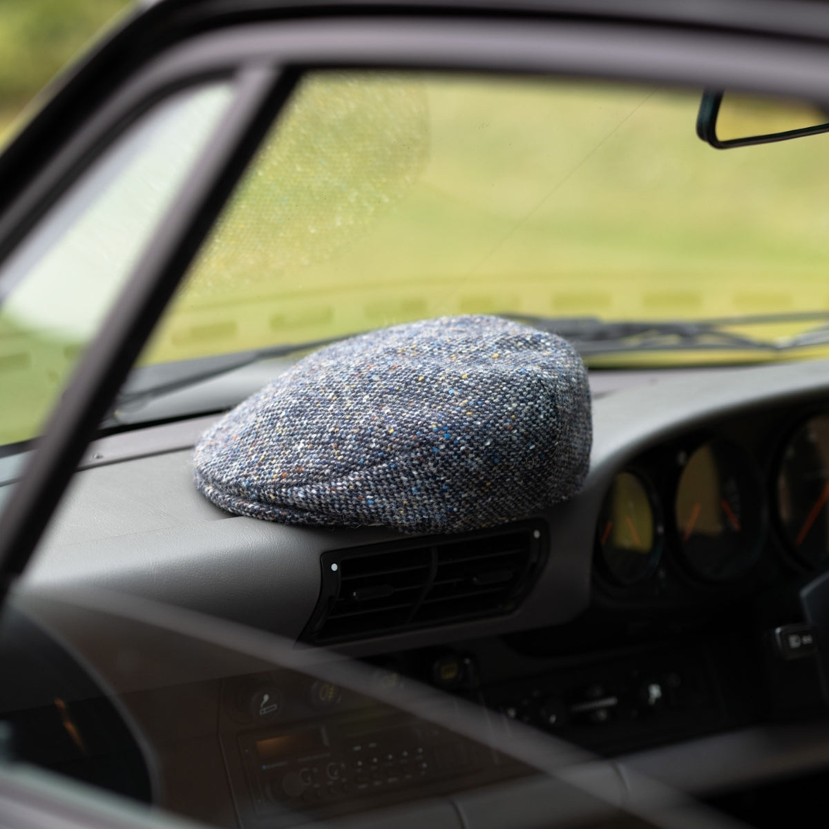 A blue tweed flat cap rests on the dashboard of a car, seen through the passenger-side window with blurred green scenery in the background.