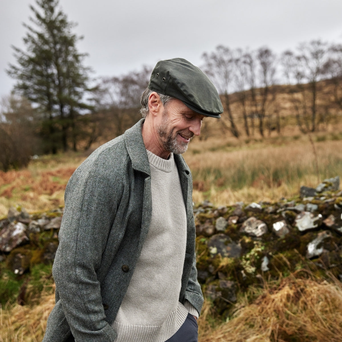 A smiling man wearing a flat cap, grey jacket, and light sweater walks outdoors by a stone wall in a grassy, rural landscape with trees and cloudy skies.