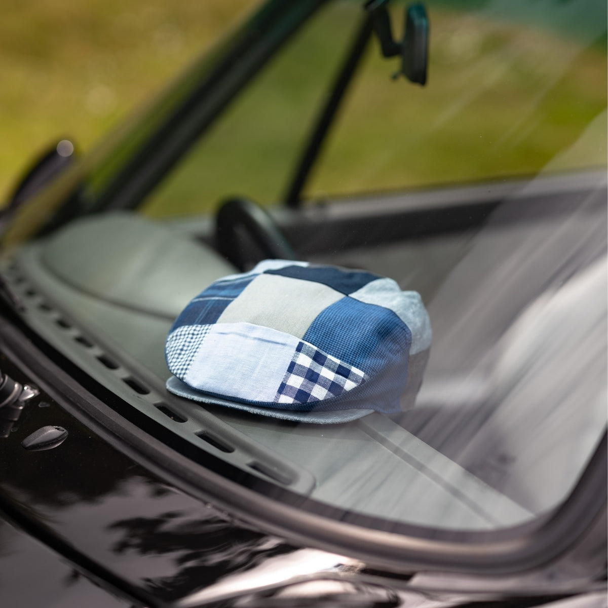 A blue patchwork flat cap rests on the dashboard of a car, visible through the windshield with blurred greenery in the background.