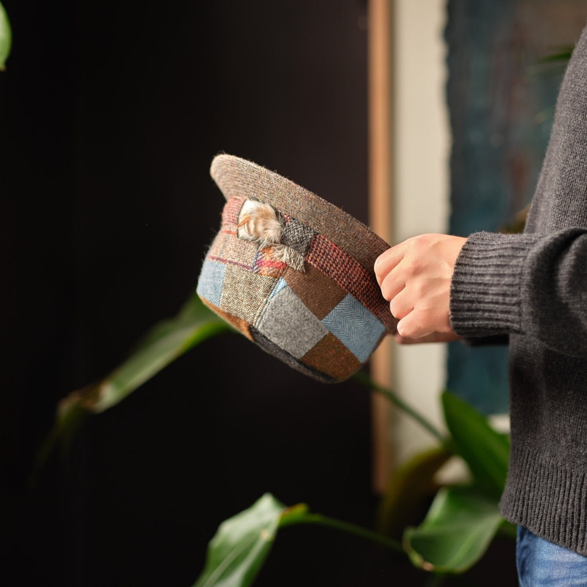 A person in a gray sweater holds a patchwork hat sideways. A small kitten peeks out of the hat, with plants and a blurred background visible behind them.