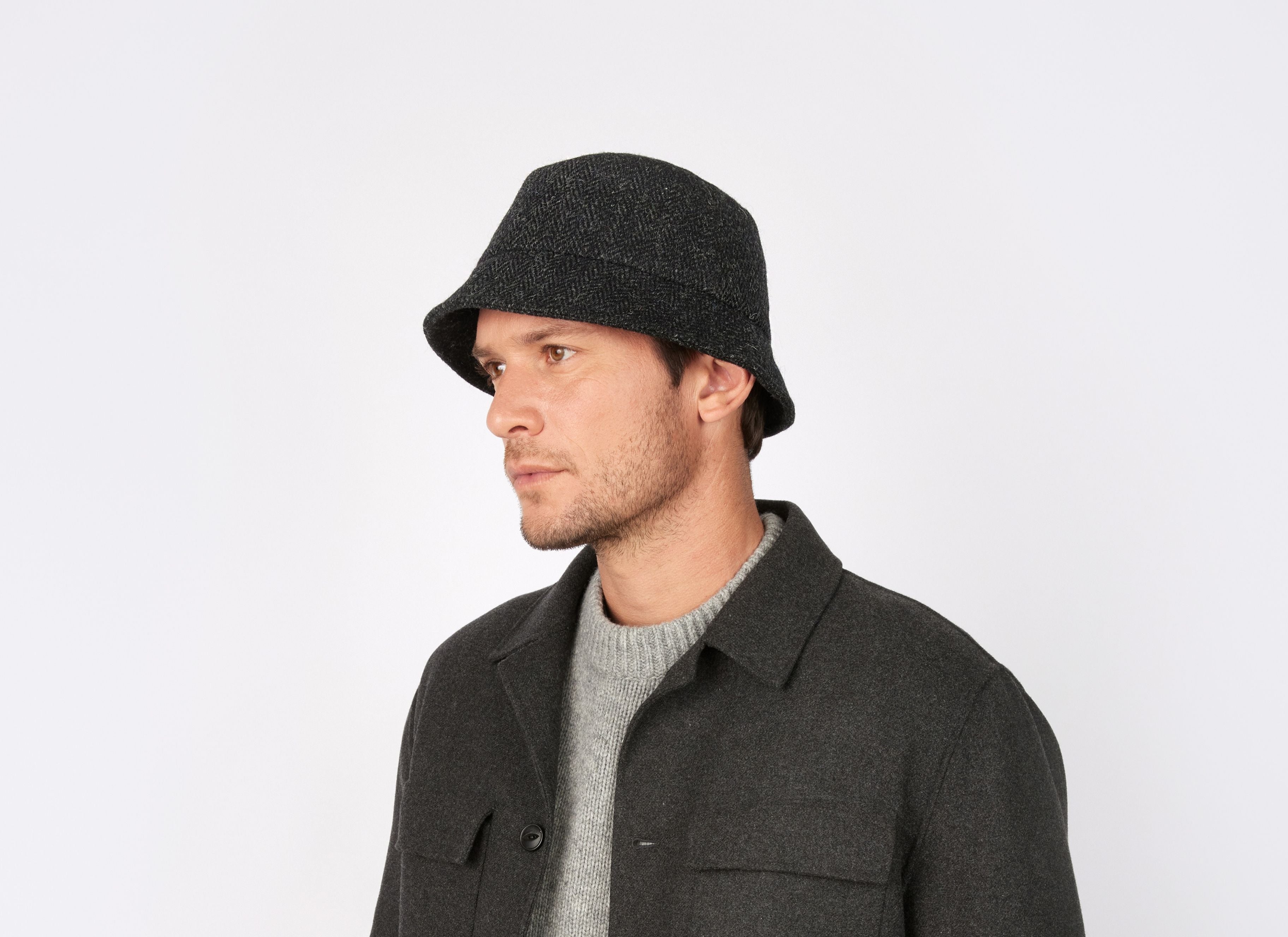 A man wearing a gray Hanna Hats of Donegal Ltd Wee Thatch Hat Tweed bucket hat, a gray sweater, and a dark gray jacket looks to the side against a plain white background.
