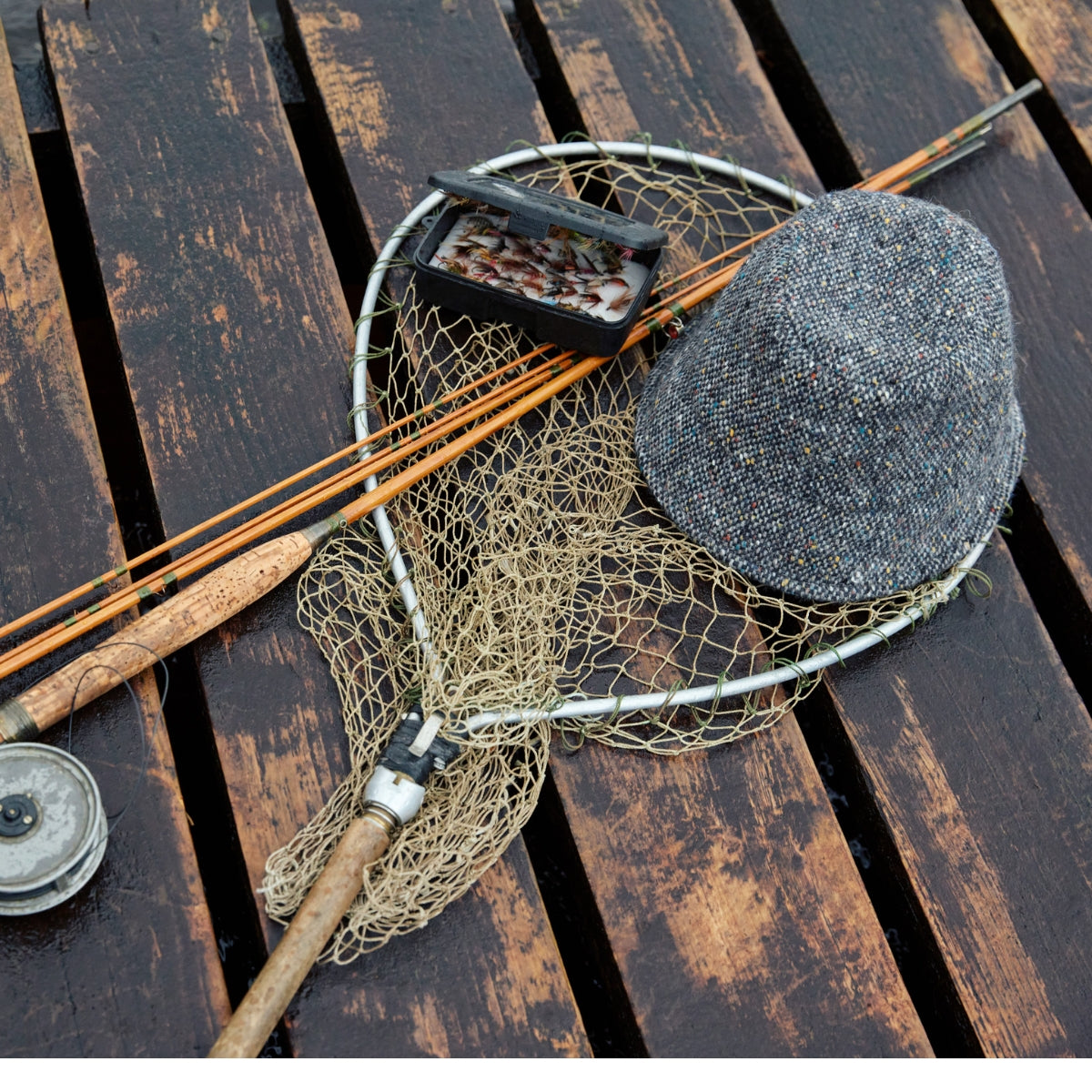 A fishing net, fishing rods, a small box of fishing flies, and a gray tweed hat are laid out on a wet wooden dock.