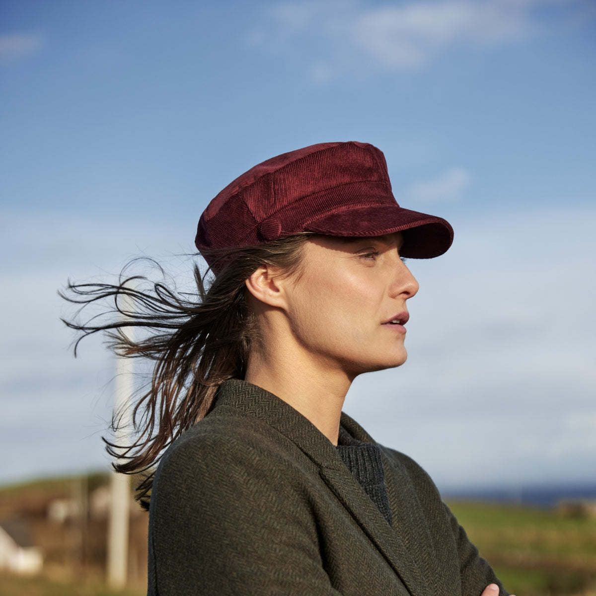 A woman wearing a dark red cap and dark jacket stands outdoors with her arms crossed, looking to the side. Her hair is blowing in the wind and the sky is blue with some clouds.