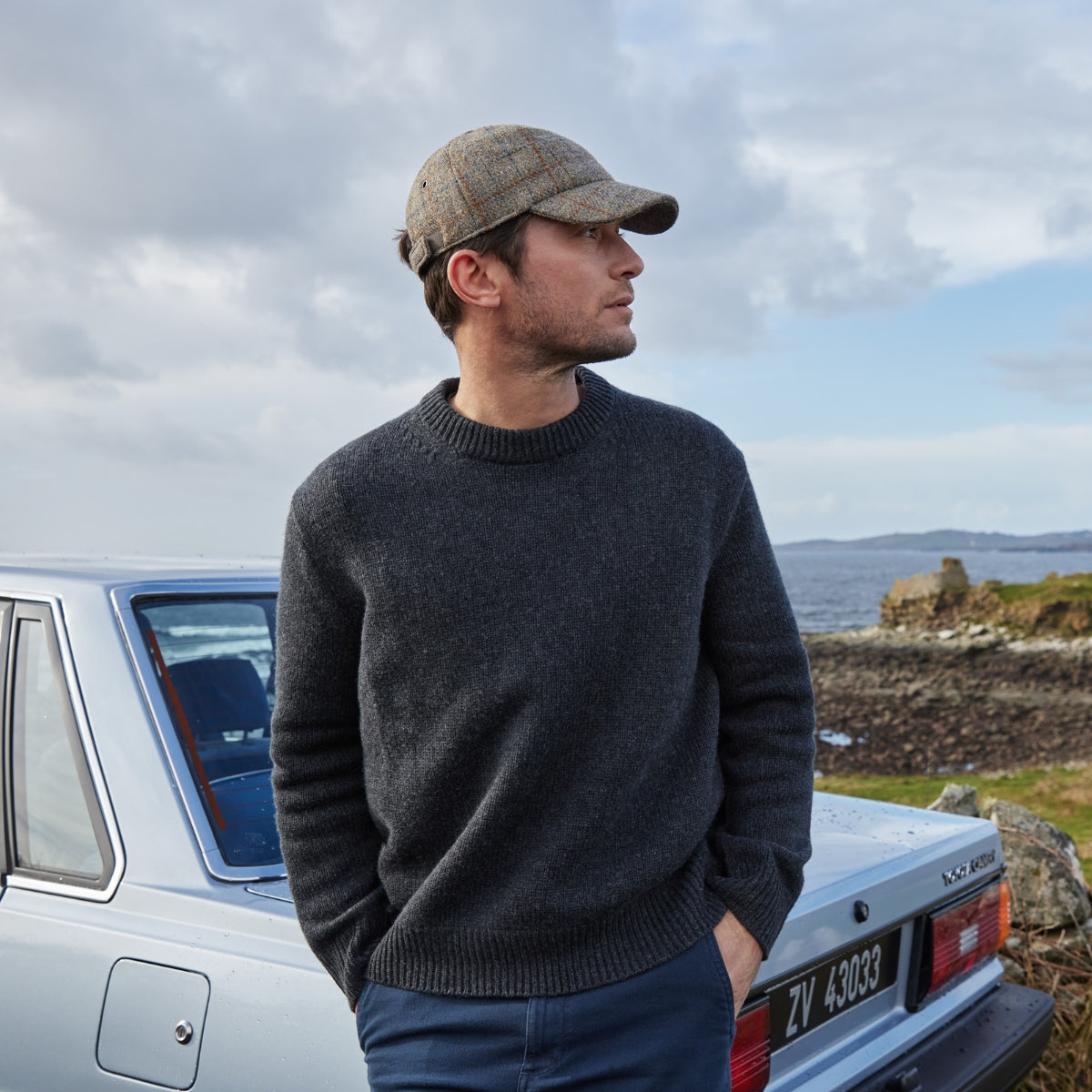 A man wearing a grey sweater and a plaid cap stands by a light blue car near a rocky coastline, looking to the side. The sky is cloudy and the sea is visible in the background.