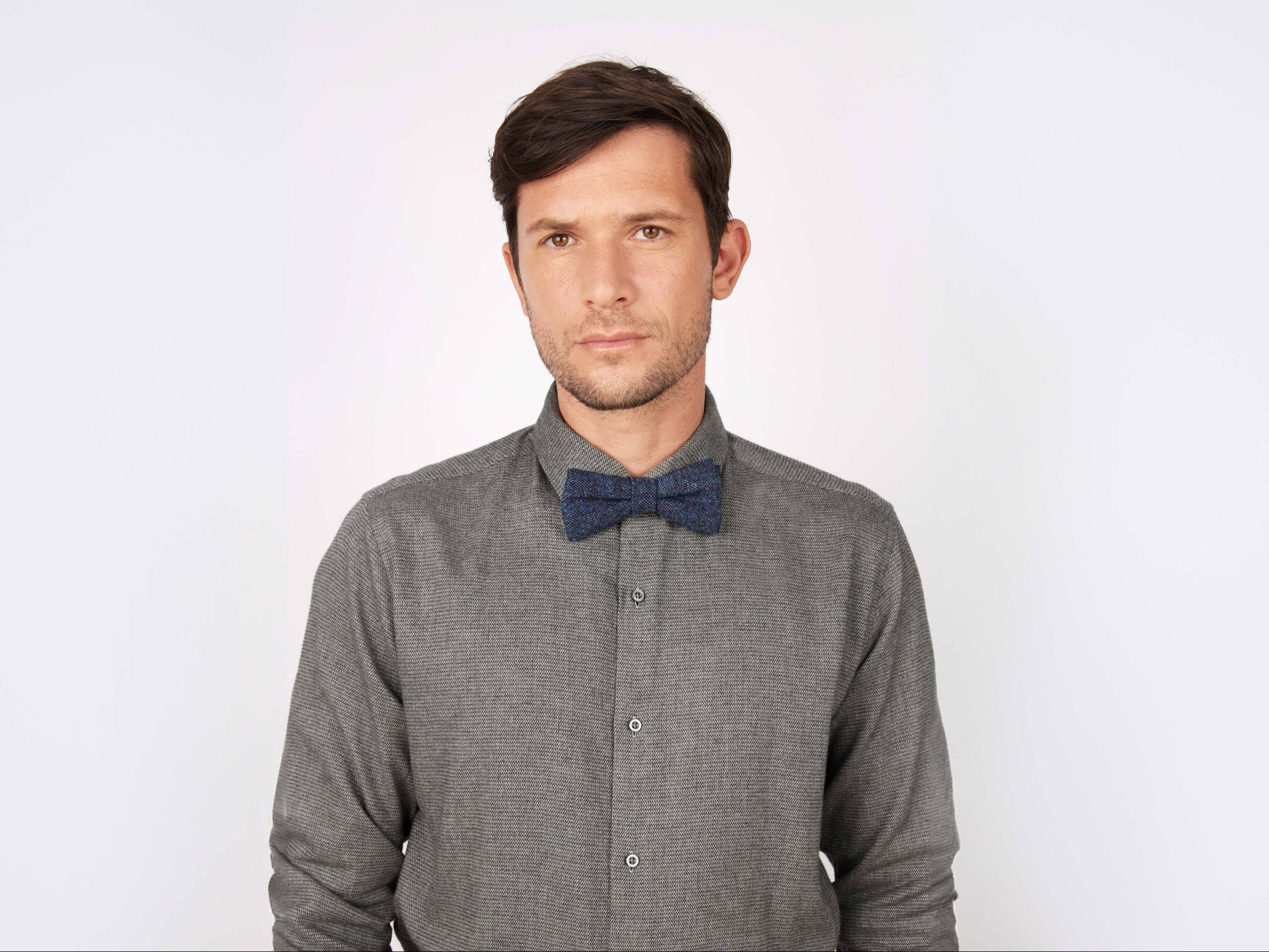 A man with short brown hair wears a gray button-up shirt and the Hanna Hats Bow Tie Tweed, standing against a plain white background and looking directly at the camera with a neutral expression.