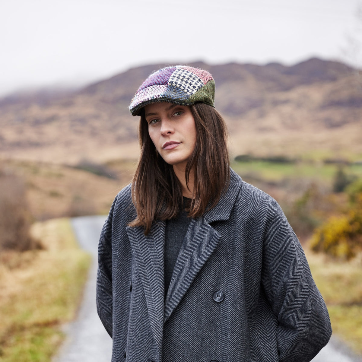 A woman with long brown hair wears a patchwork cap and a gray herringbone coat, standing on a country road with rolling hills and cloudy skies in the background.