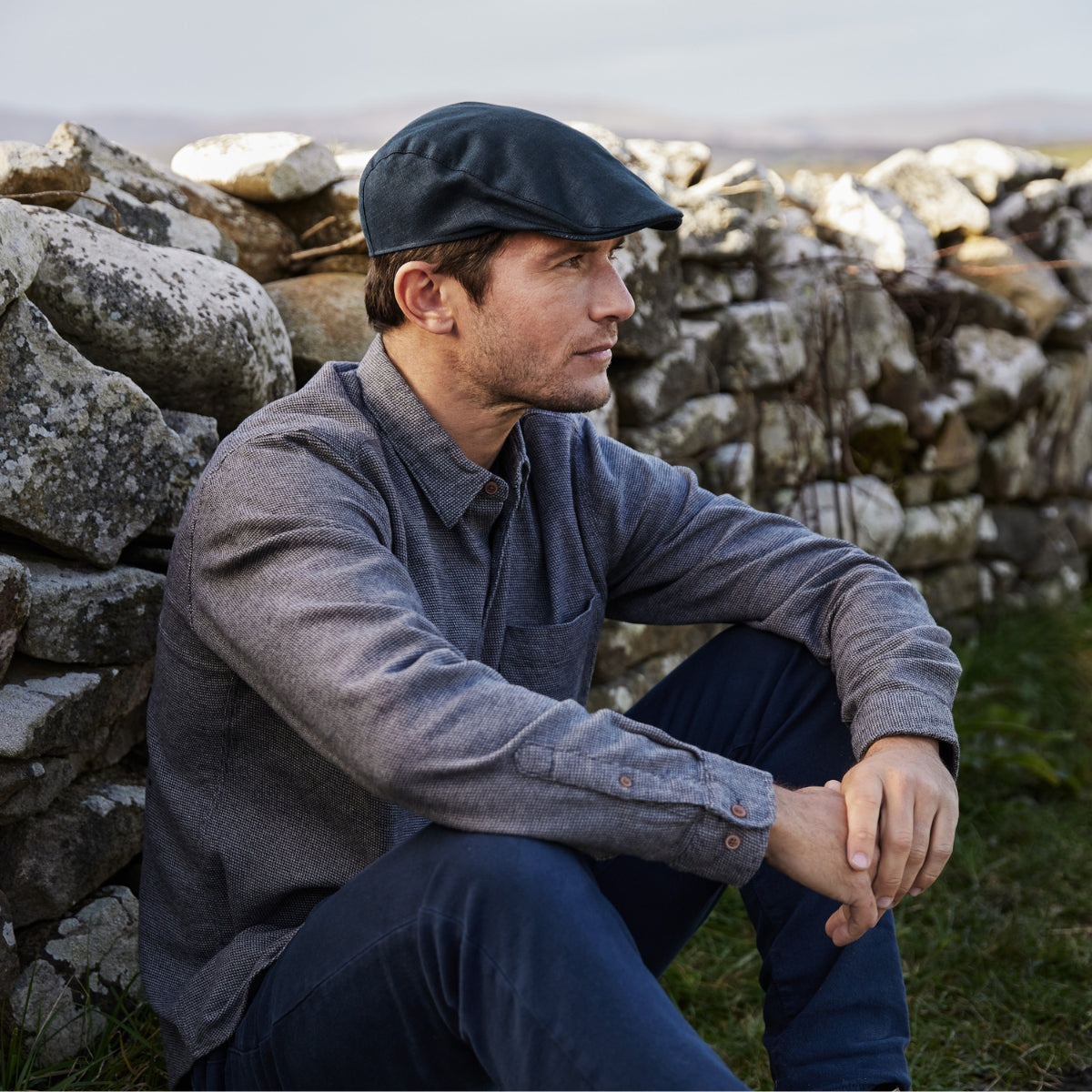 A man wearing a flat cap and gray shirt sits outdoors against a stone wall, looking thoughtfully into the distance. The background features grass and a blurred natural landscape.
