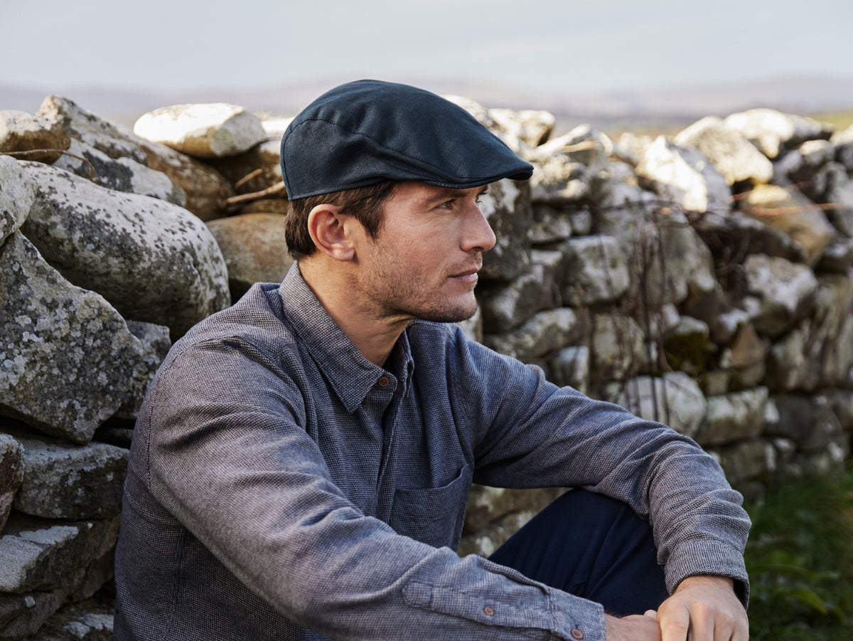 A man wearing a flat cap and gray shirt sits outdoors against a stone wall, looking thoughtfully into the distance. The background features grass and a blurred natural landscape.