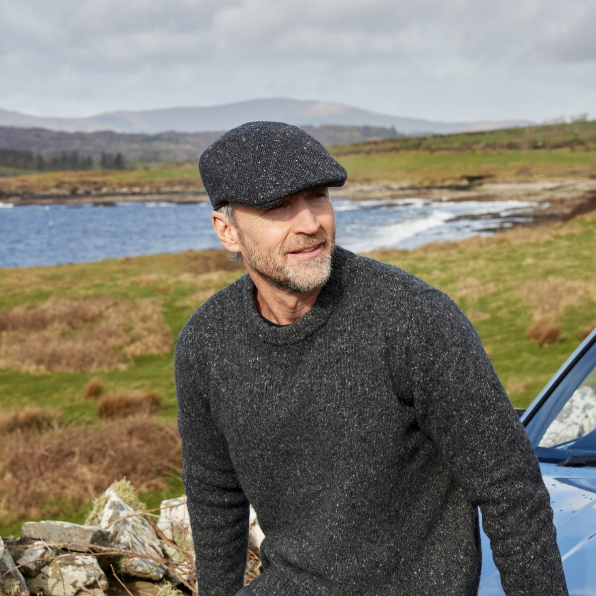 A man wearing a dark flat cap and a gray sweater sits outdoors near a lake and grassy fields, with hills in the background and a blue car nearby.
