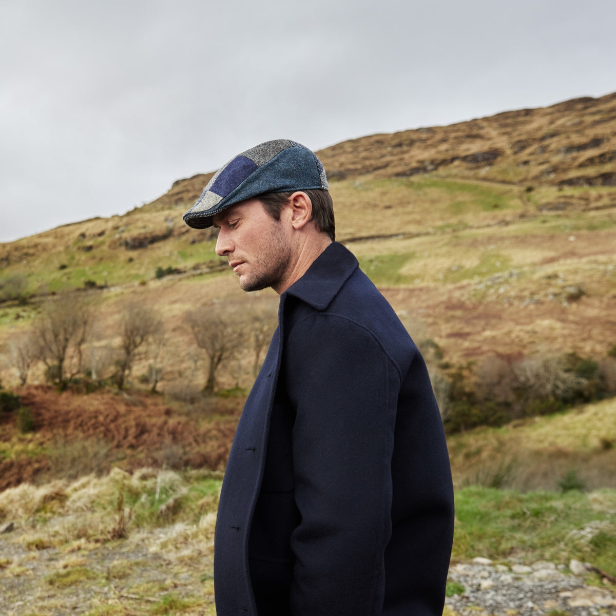 A man wearing a dark coat and a plaid flat cap stands in profile outdoors, with his eyes closed, against a backdrop of grassy hills and a cloudy sky.