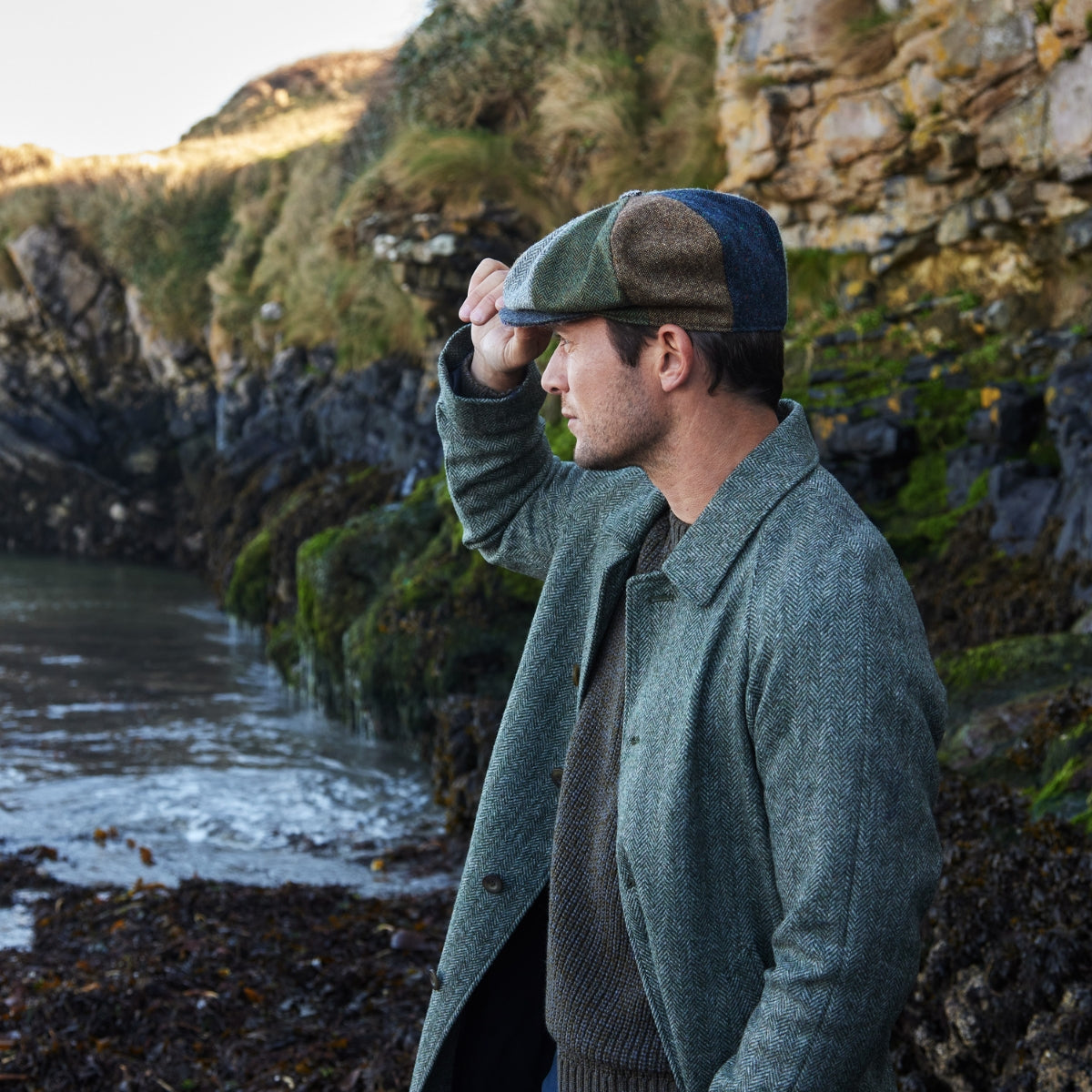 A man in a green tweed coat and patchwork flat cap stands by a rocky shoreline, holding the brim of his cap, with cliffs and mossy rocks in the background.