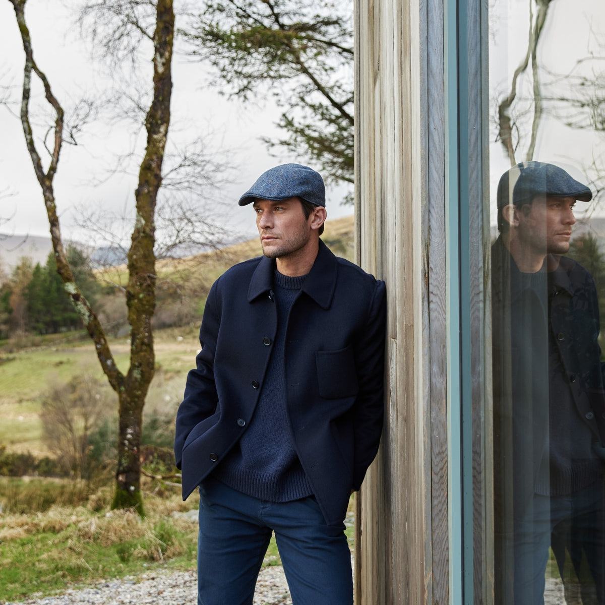 A man in a navy coat, sweater, and flat cap leans against a wooden building with large glass windows, reflecting his image. Leafless trees and grassy hills are visible in the background under an overcast sky.