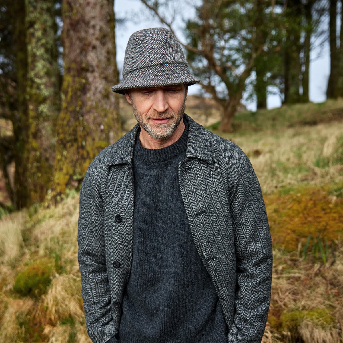 A man wearing a gray tweed jacket, dark sweater, and tweed hat stands outdoors in a grassy, wooded area with trees and mossy ground.