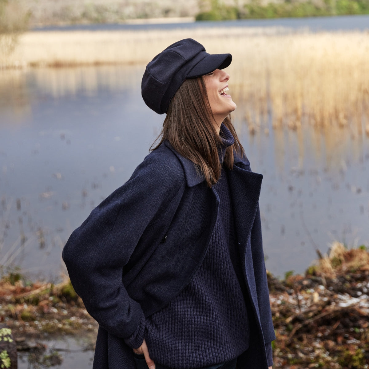 A woman wearing a navy coat, sweater, and cap stands outdoors near a lake, laughing and looking to the side. Tall grasses and water are visible in the background.