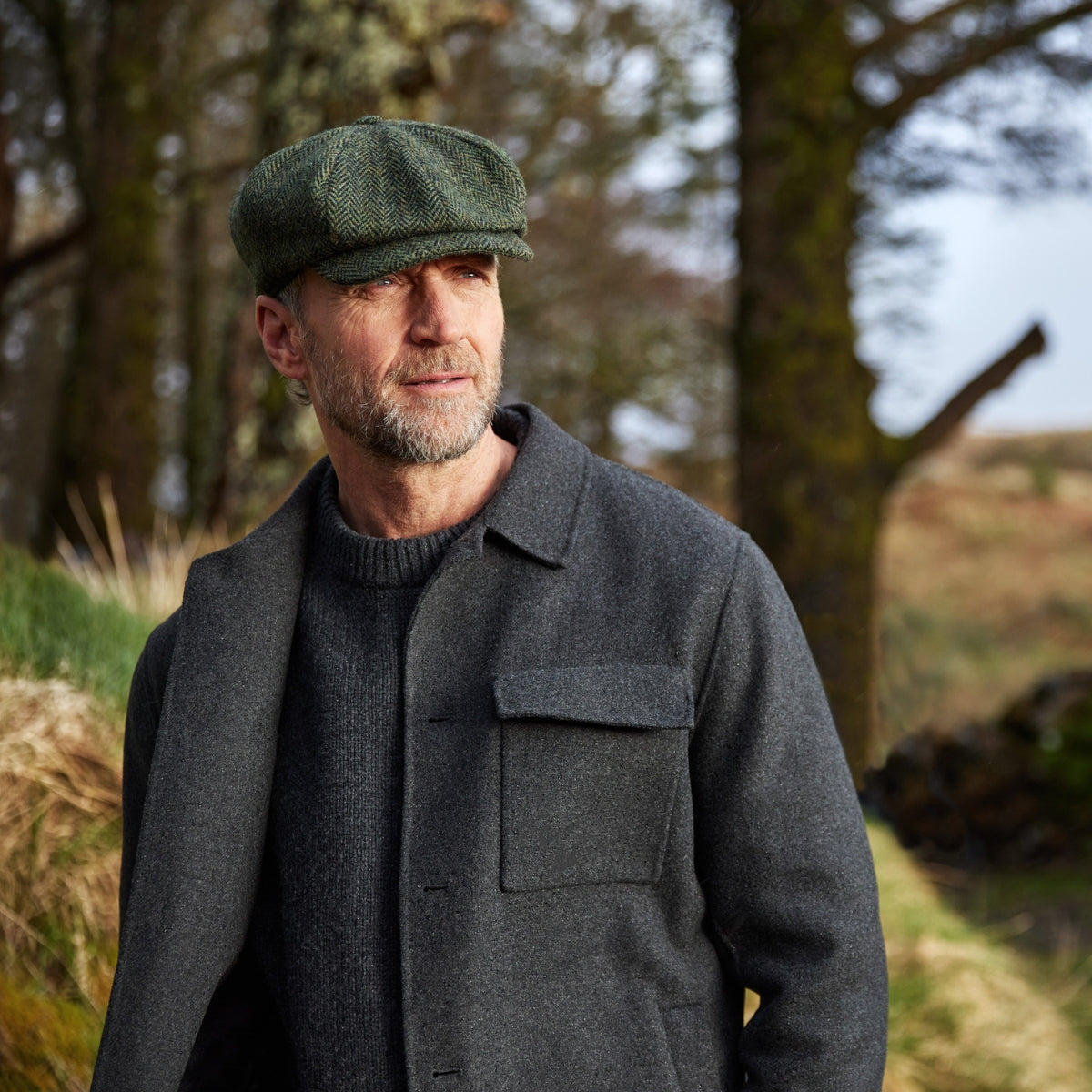 A middle-aged man with a beard, wearing a green flat cap and a dark coat, stands outdoors in a natural setting with trees and grass in the background.
