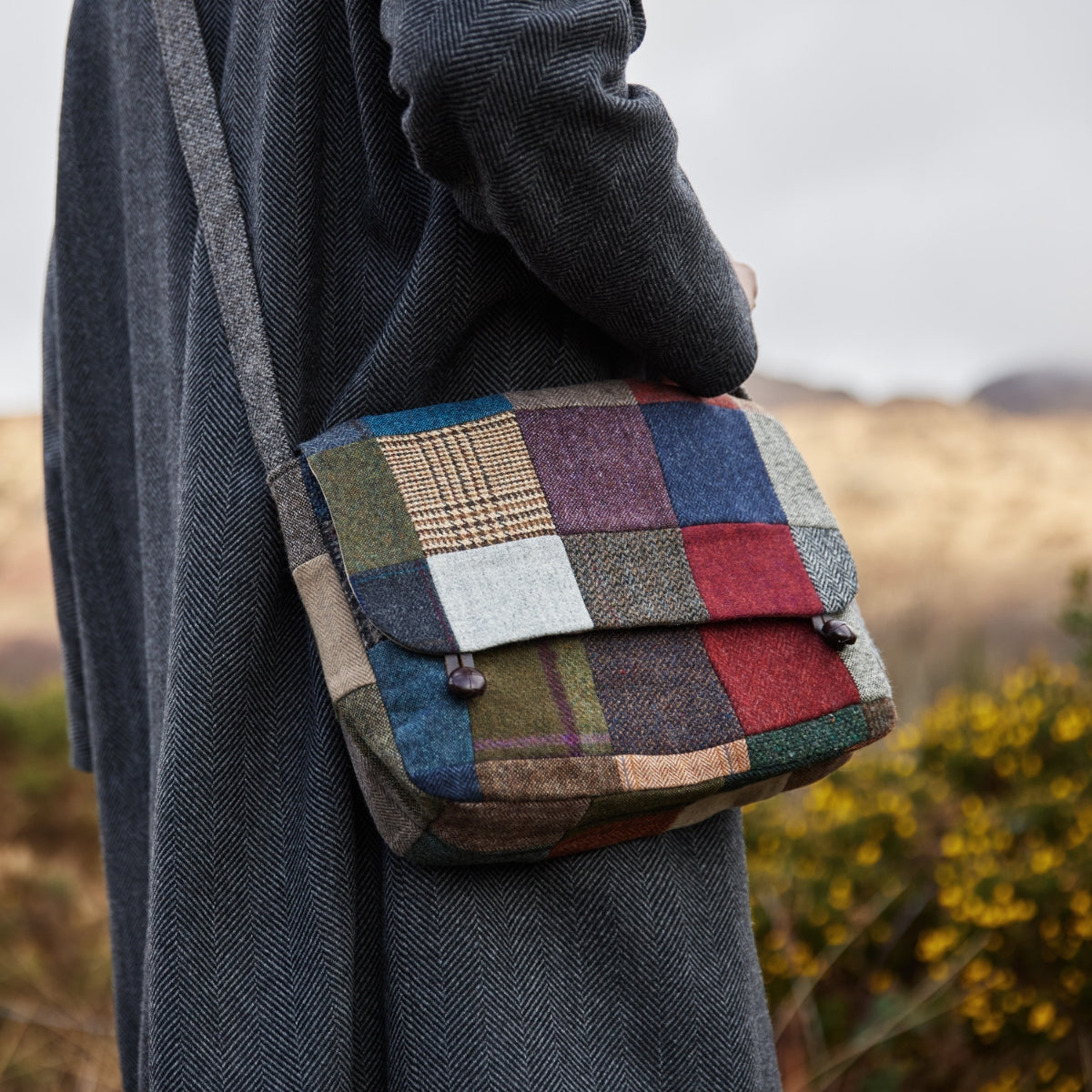 A person wearing a dark herringbone coat carries a colorful patchwork shoulder bag outdoors, with blurred greenery and hills in the background.