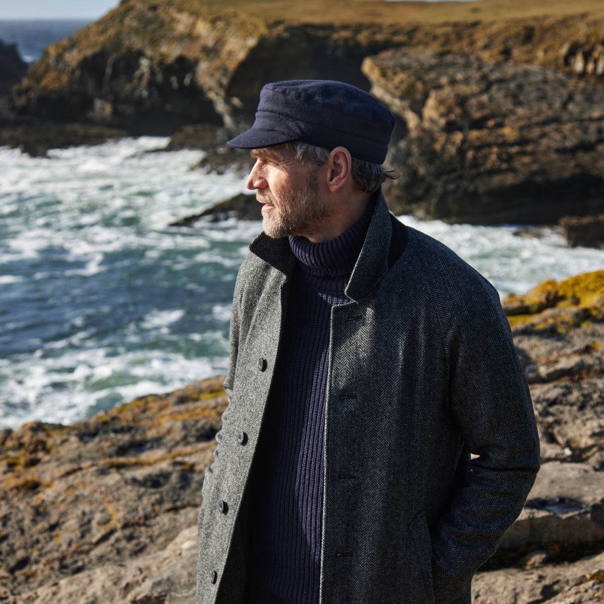 A man in a gray coat and navy cap stands on rocky cliffs by the ocean, looking into the distance with waves crashing behind him under a clear sky.