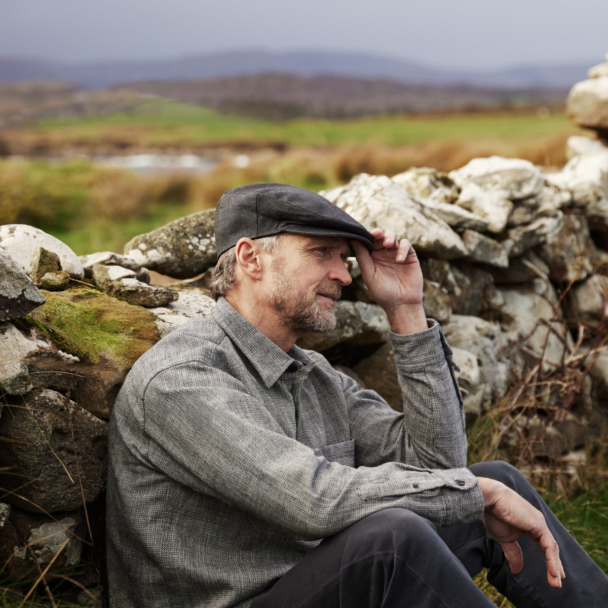 An older man wearing a flat cap and gray shirt sits outdoors, leaning against a stone wall with green fields and hills in the background. He looks thoughtful, holding the brim of his hat.