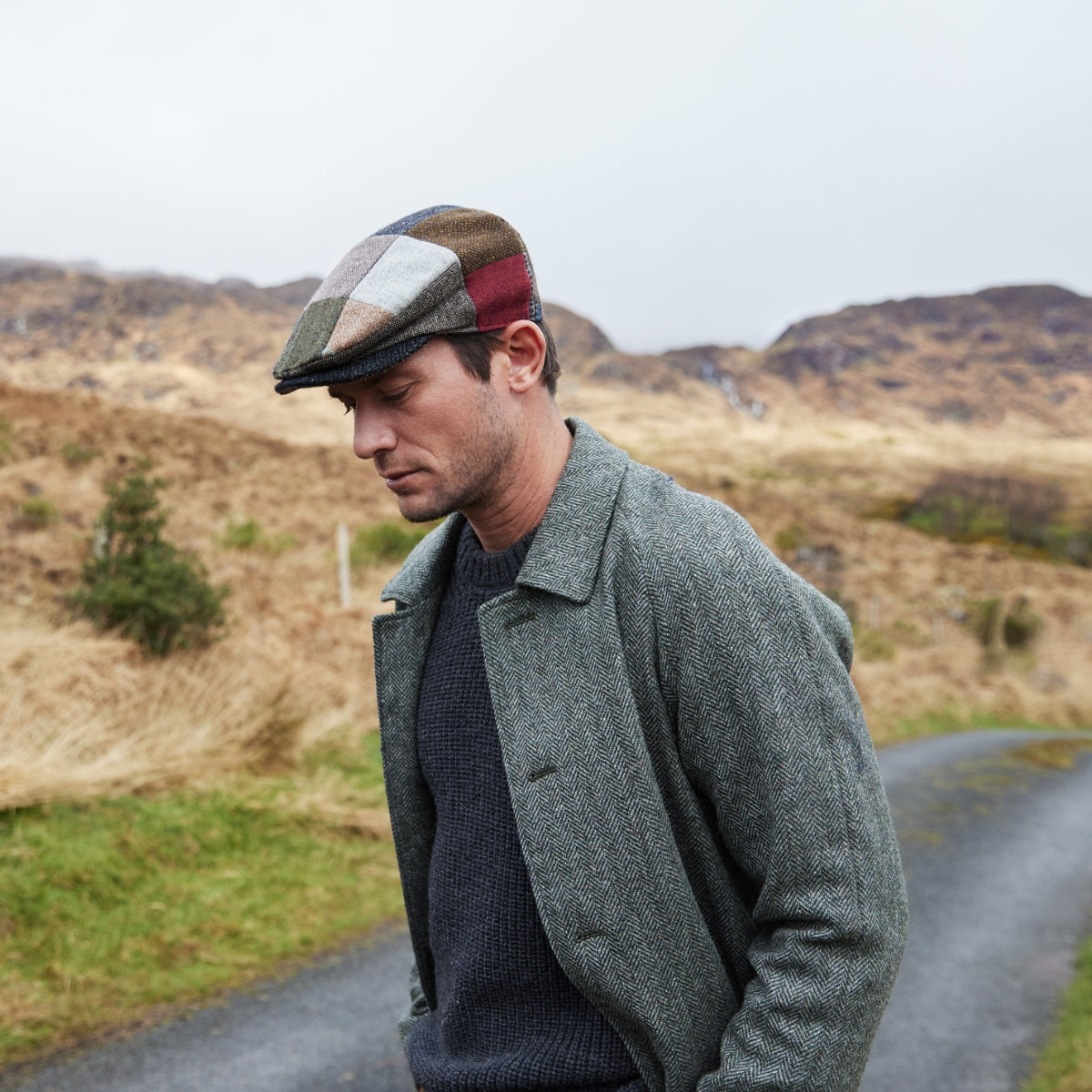A man wearing a patchwork flat cap, gray coat, and dark sweater walks outdoors on a rural road, with grassy hills and cloudy sky in the background.