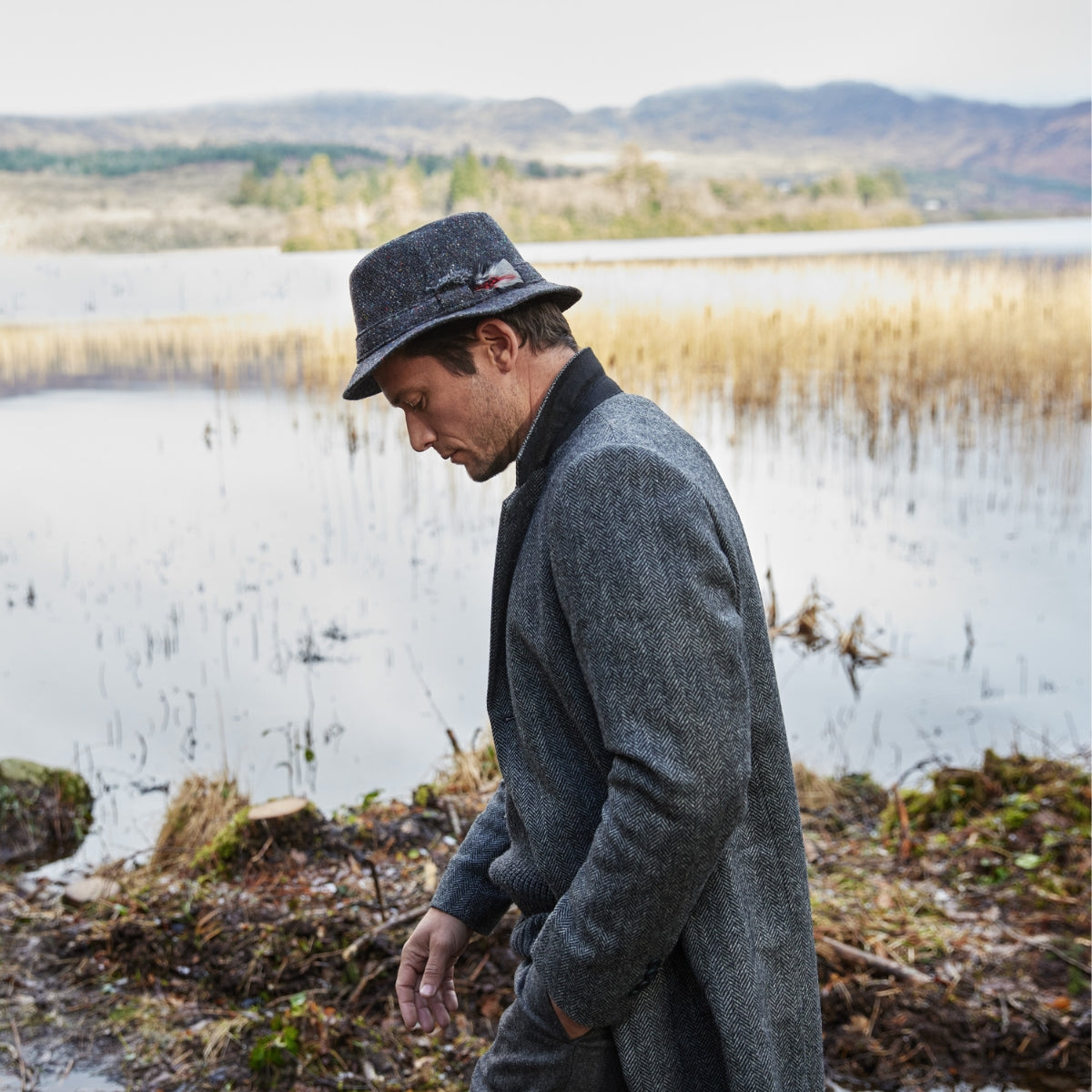 A man in a gray coat and hat walks along a lakeshore surrounded by reeds and autumn foliage, with hills and cloudy sky in the background.