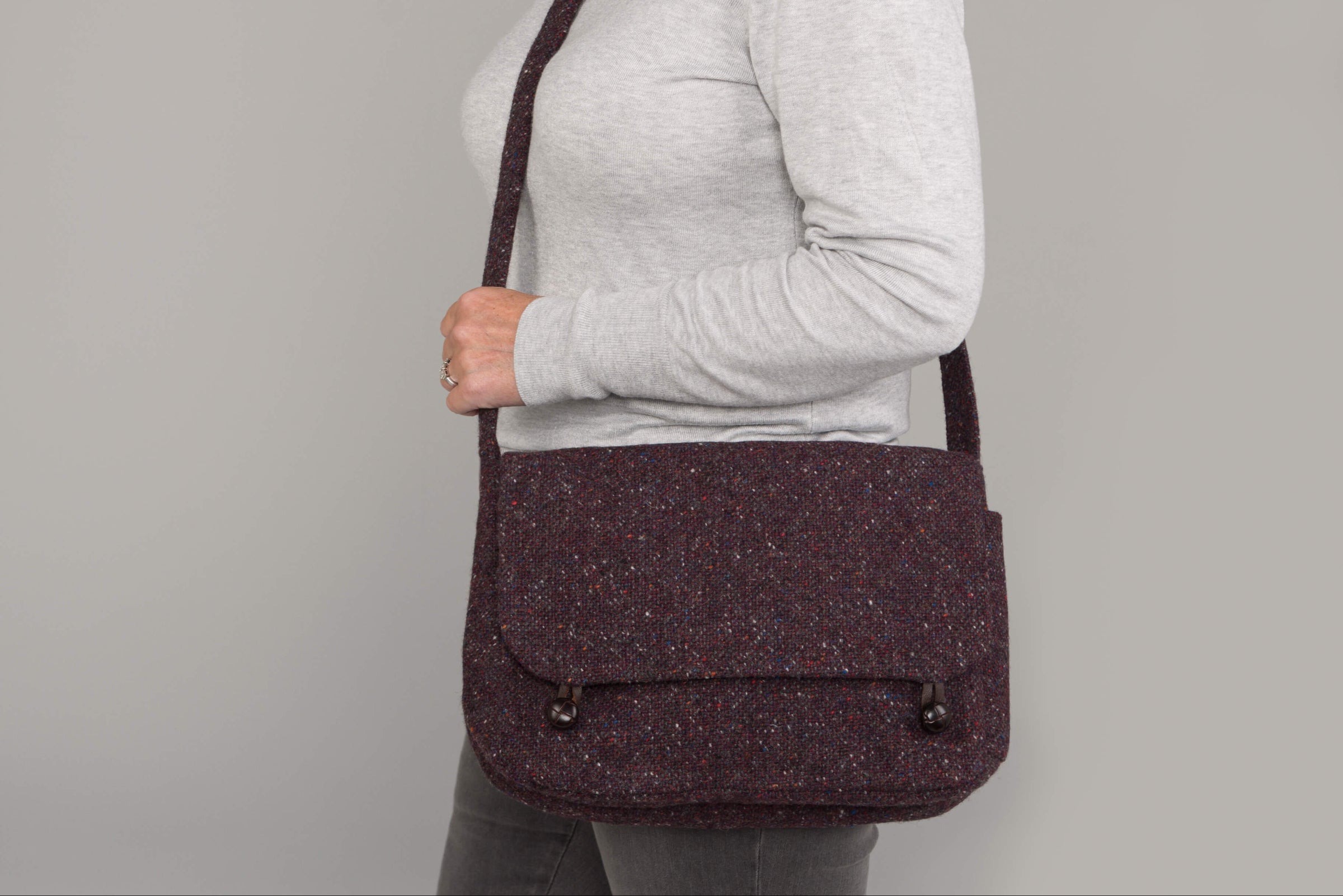 A person in a light grey long-sleeve shirt and dark pants carries the Hanna Hats Satchel Bag Tweed, a dark textured everyday satchel with flap and button closures. The plain, neutral background highlights the accessory.