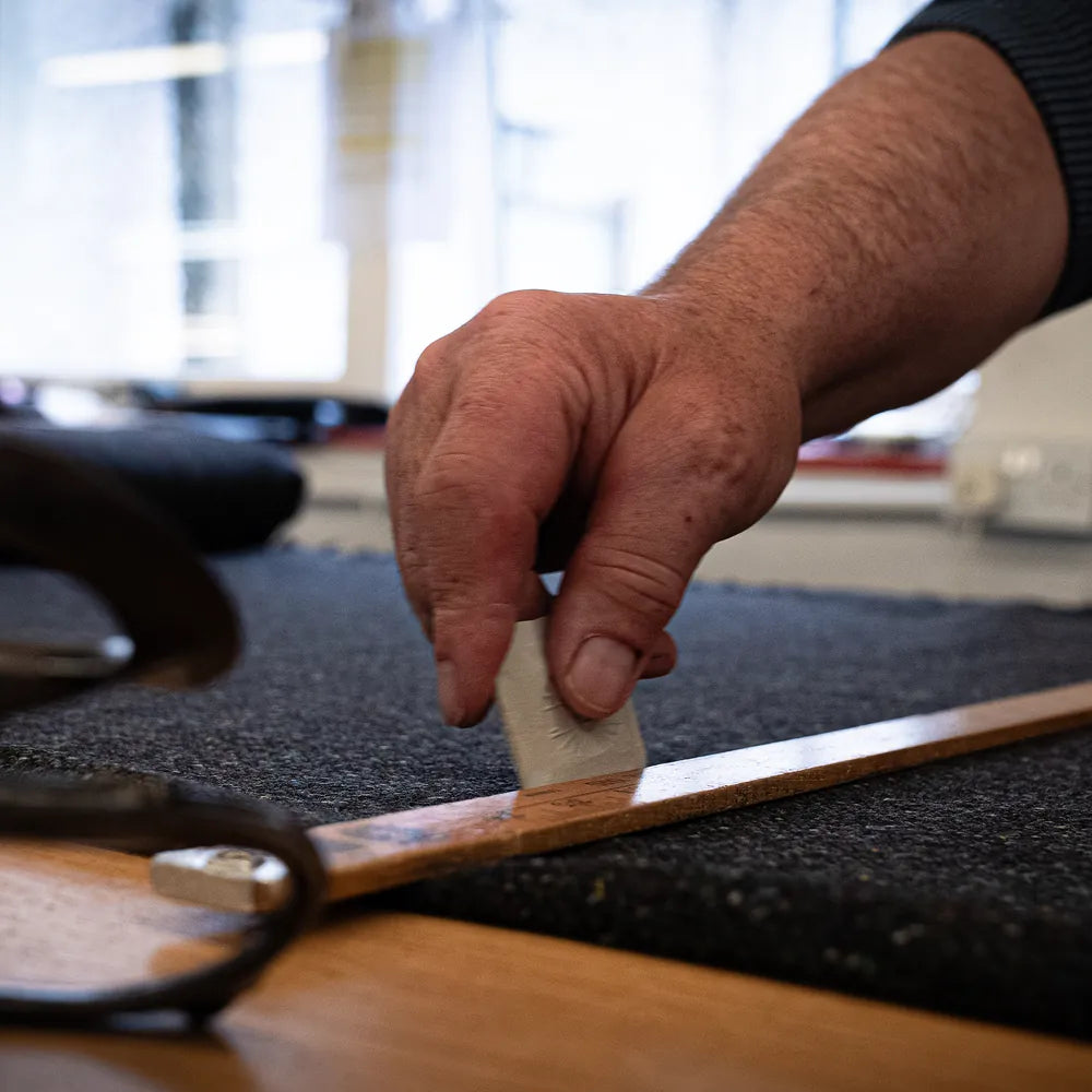 A close-up of a person’s hand holding a piece of tailor’s chalk, marking a measurement on a strip of fabric or wood on a worktable, with blurred tools and background.