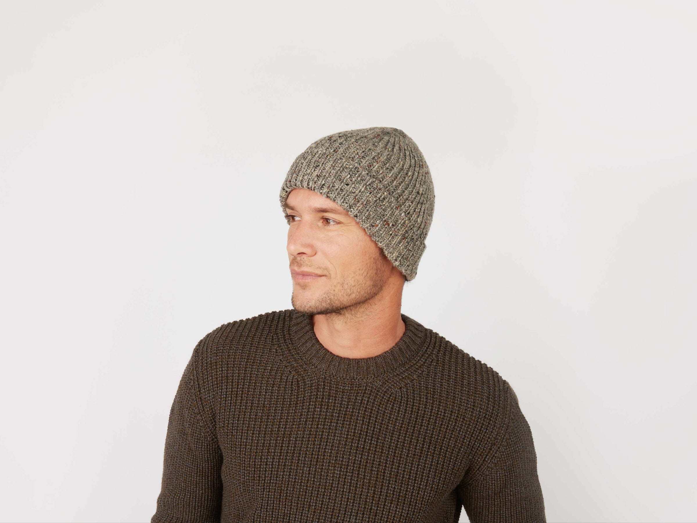 A man wearing a gray Donegal Beanie by Hanna Hats of Donegal Ltd and a brown textured sweater looks to his left against a plain white background.