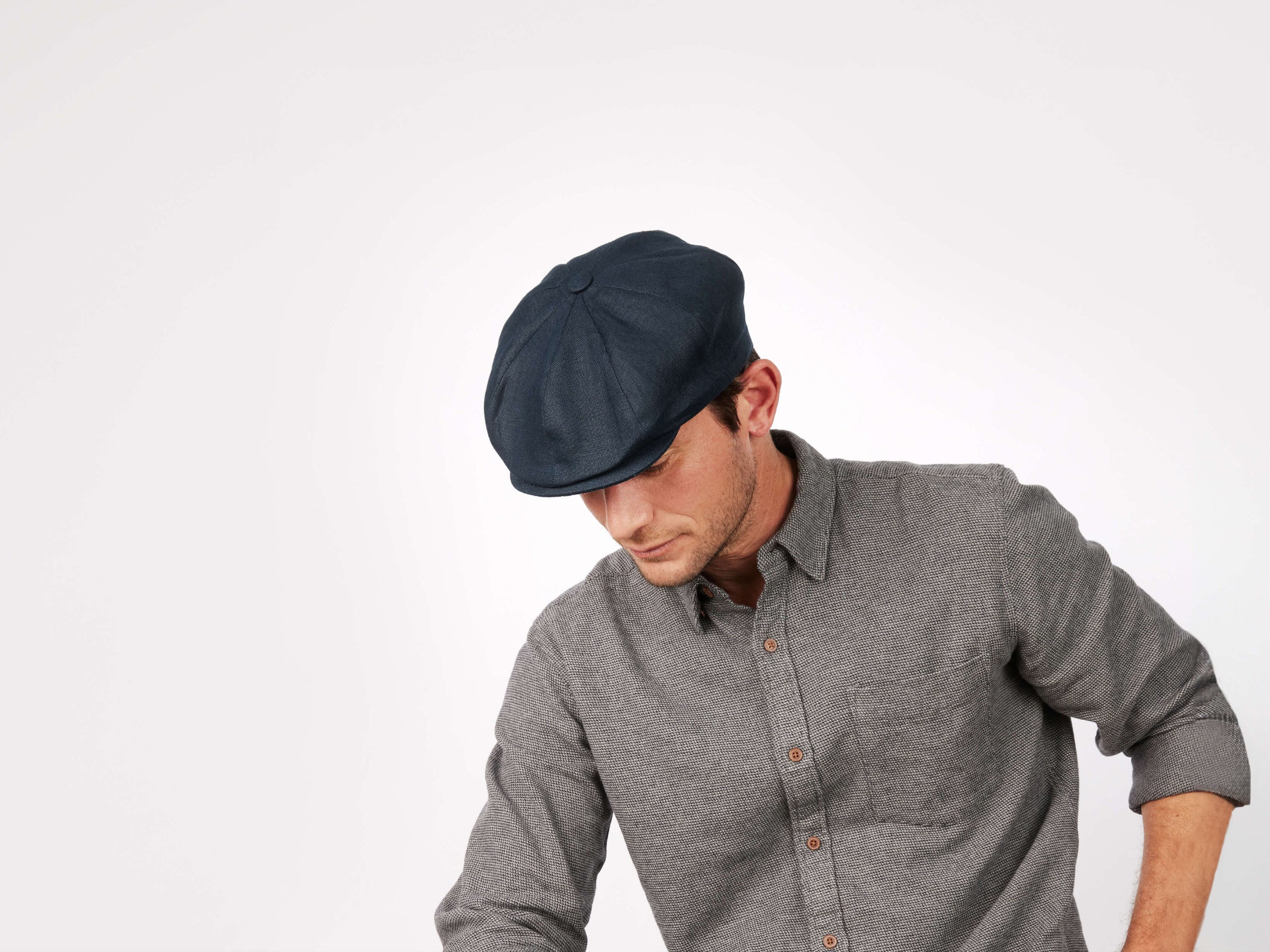 A man in a dark Connery Cap Linen by Hanna Hats, made from recycled plastic, and a grey button-up shirt looks downward against a plain white background.