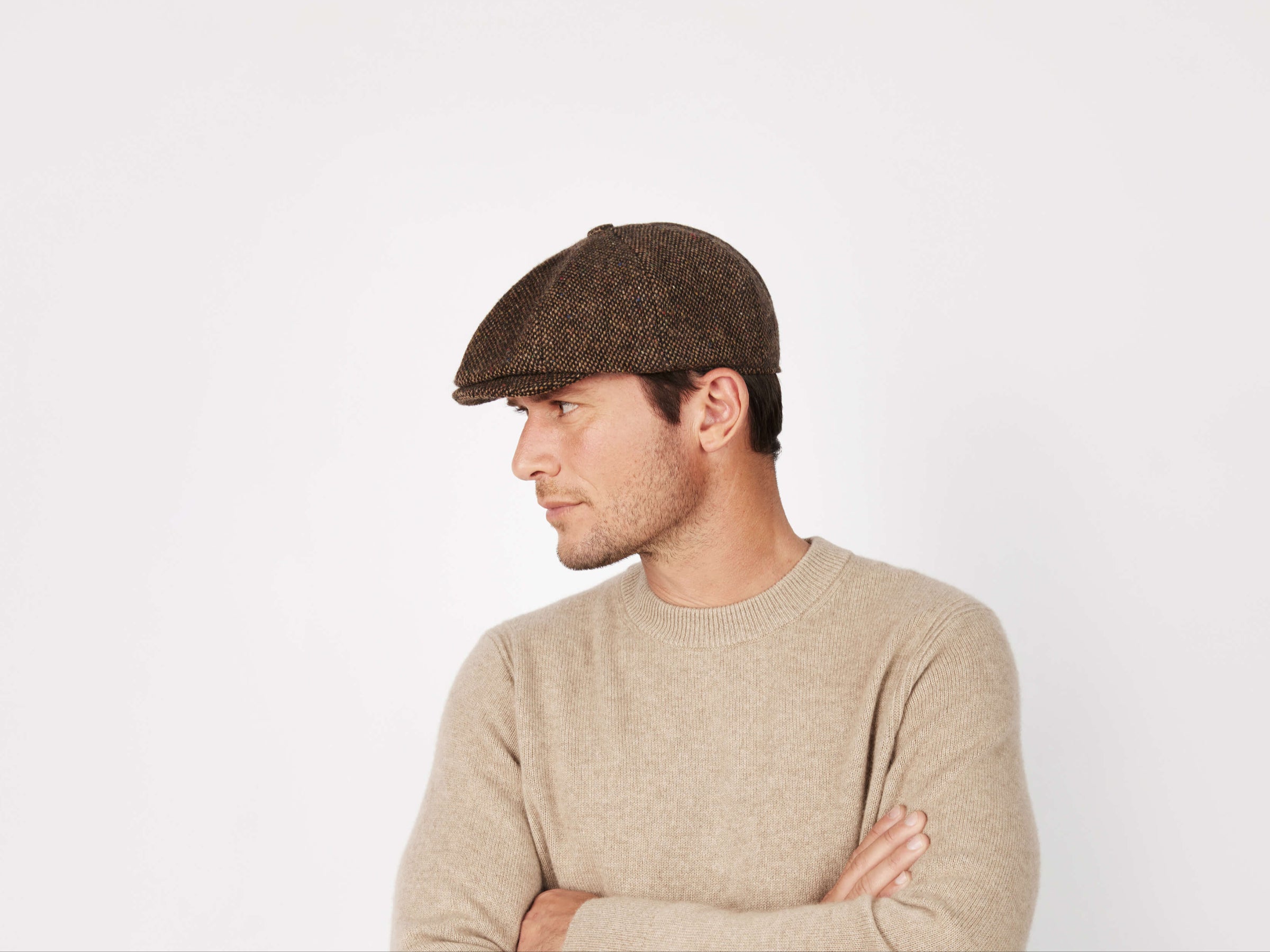 A man wears a Hanna Hats Connery Cap Tweed and a beige sweater, standing with arms crossed and looking left against a plain white background.