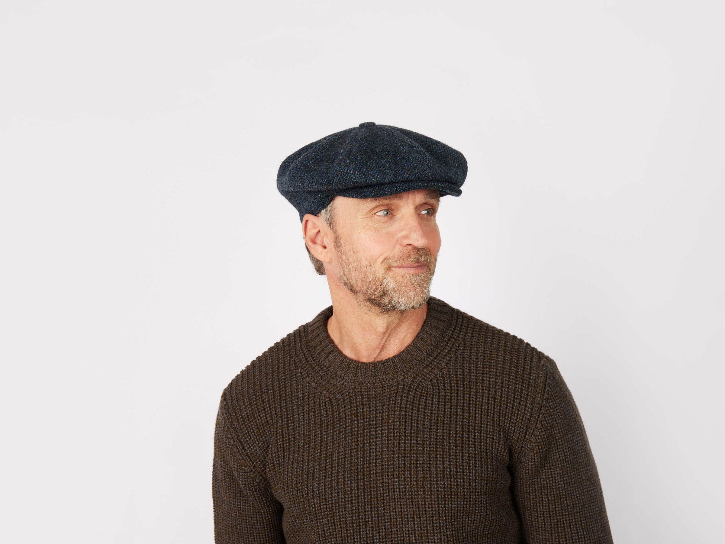 A middle-aged man with a short beard wears a Hanna Hats Eight Piece Cap Tweed and a brown textured sweater, gazing slightly left against a plain white background.