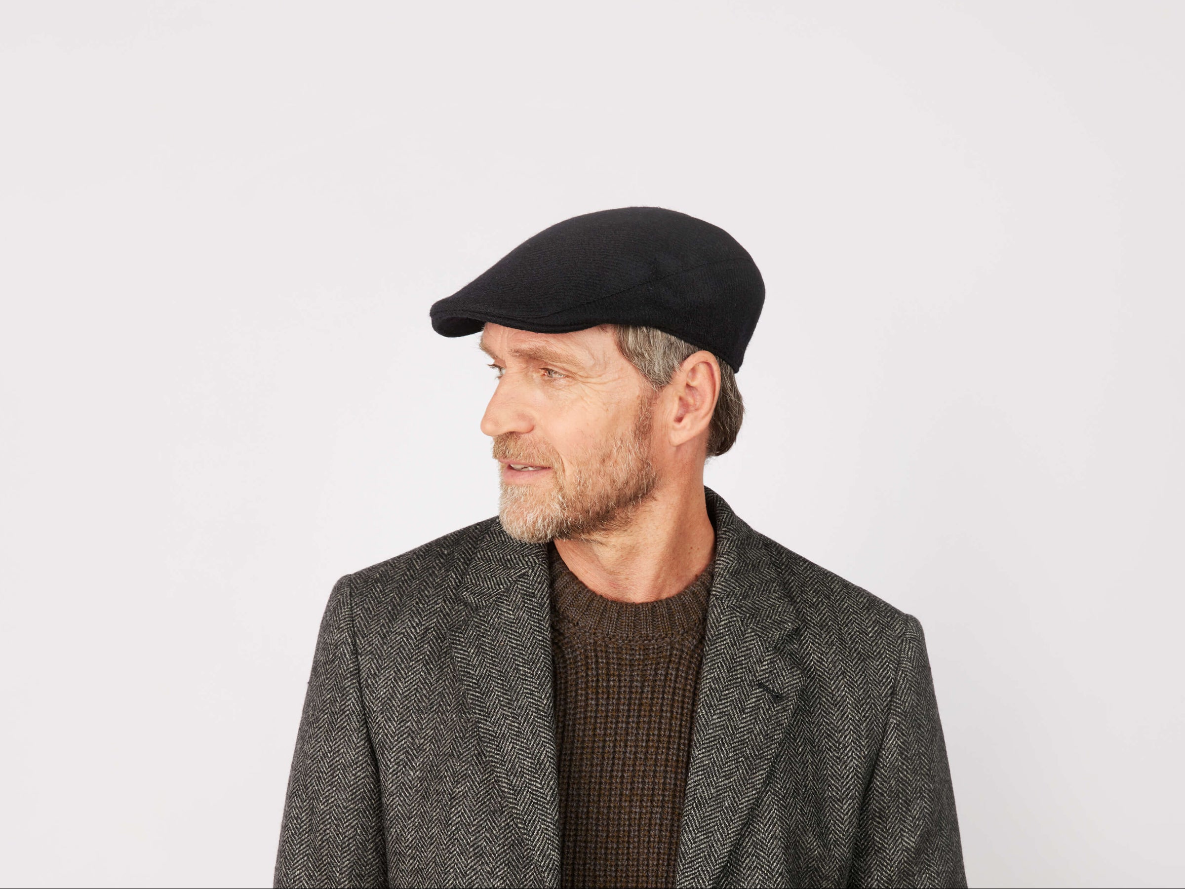 A middle-aged bearded man in a dark Hanna Hats Donegal Touring Cap Tweed, brown textured sweater, and tailored gray herringbone blazer looks to his left against a plain white background.
