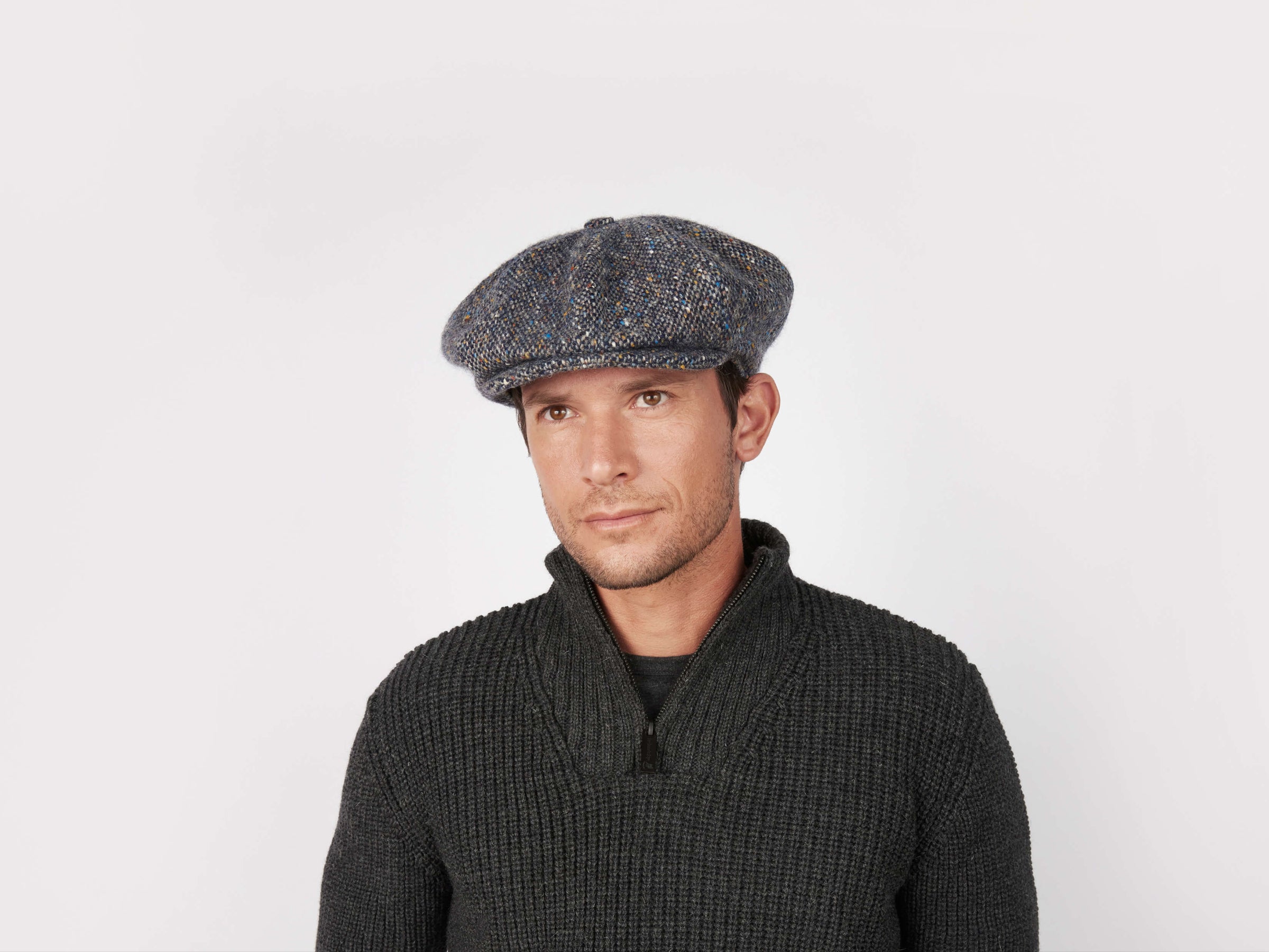 A man in a dark textured sweater and a voluminous Hanna Hats Eight Piece Cap Tweed stands against a plain white background, gazing slightly left with a neutral expression.