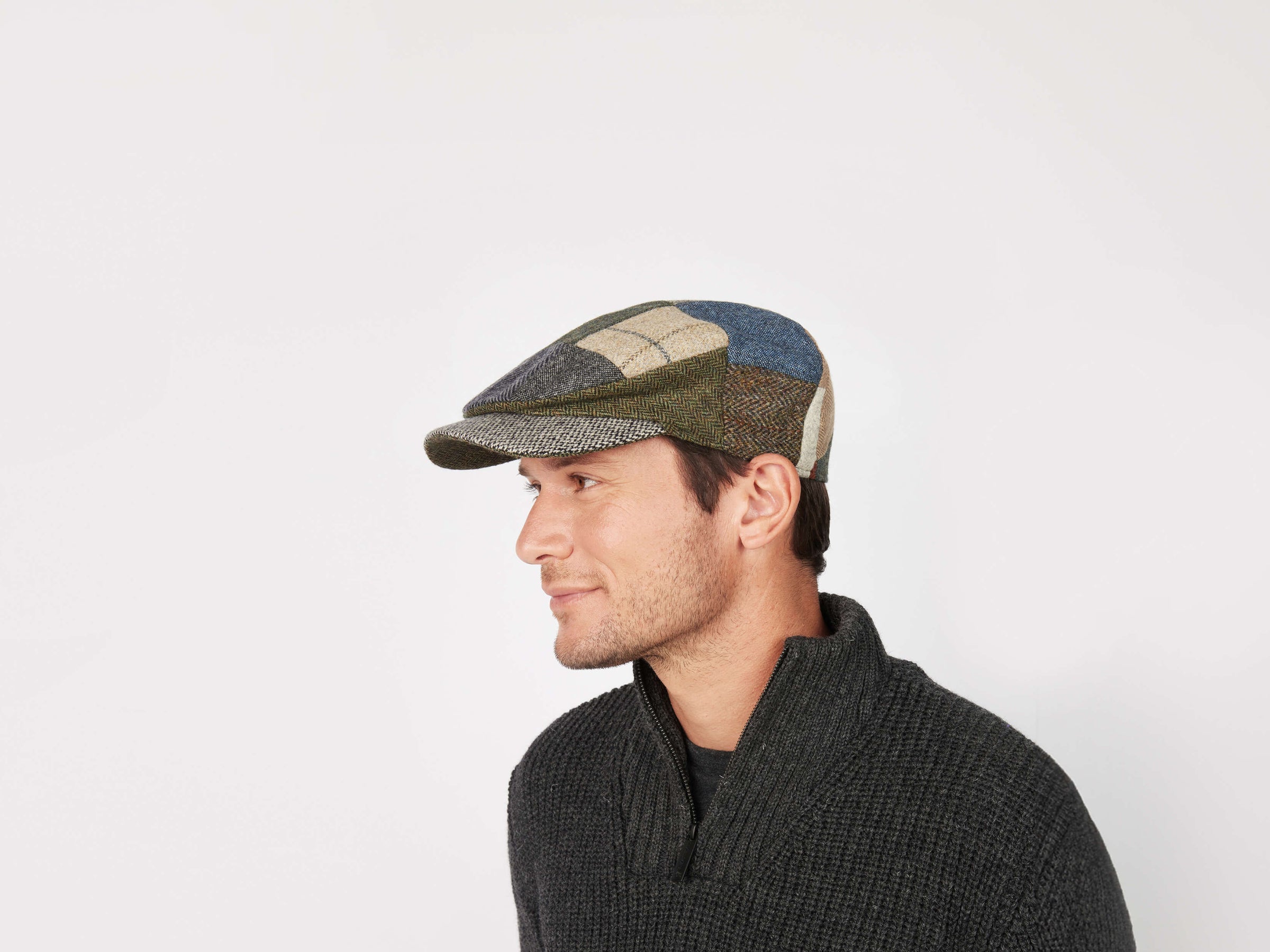 A man in a dark textured sweater and a multicolored Daithi Cap Patchwork Tweed by Hanna Hats looks left against a plain white background, highlighting the vibrant style of this Irish tweed cap.
