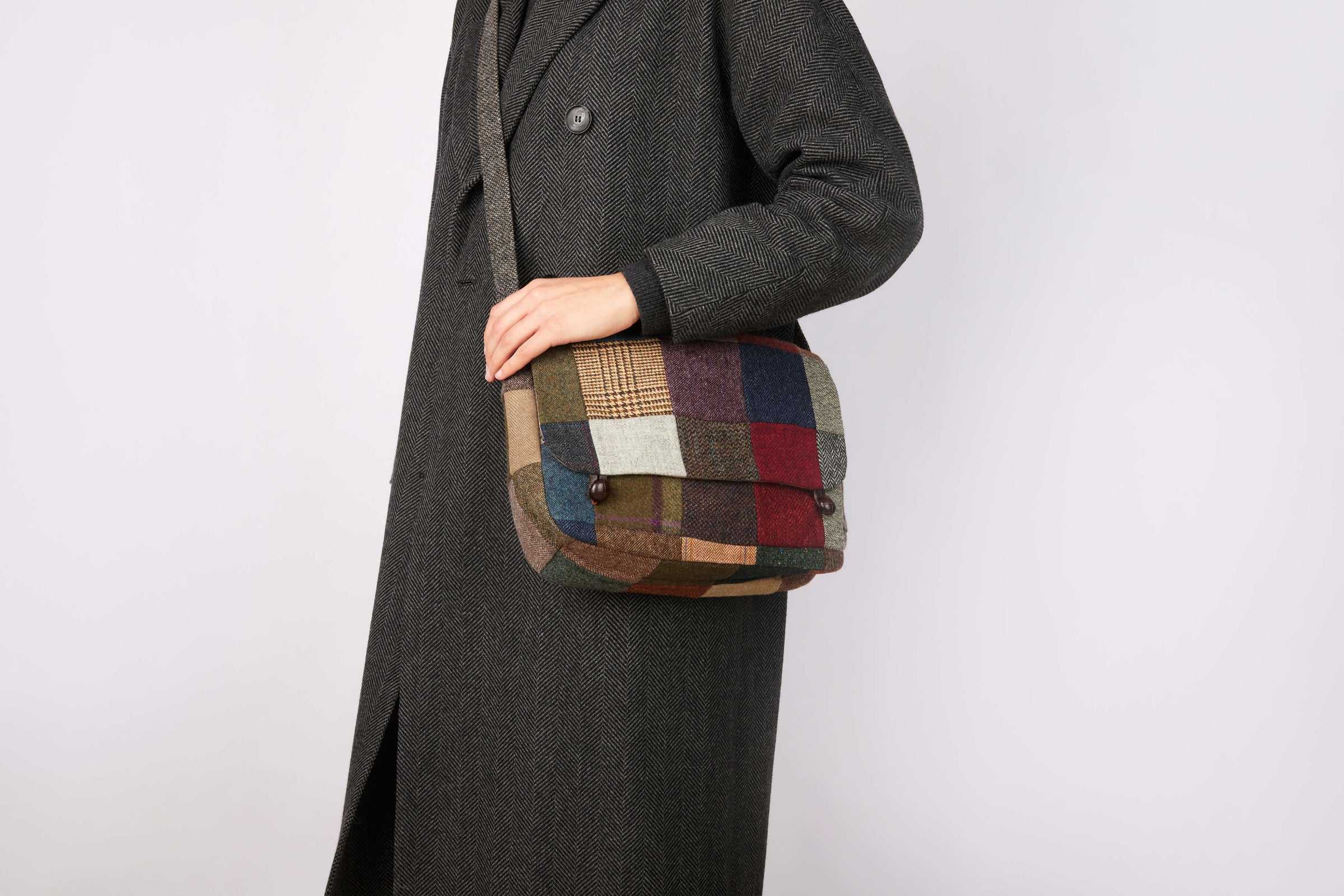 A person in a long, dark herringbone coat holds the Hanna Hats Satchel Bag Patchwork Tweed against a plain light background.