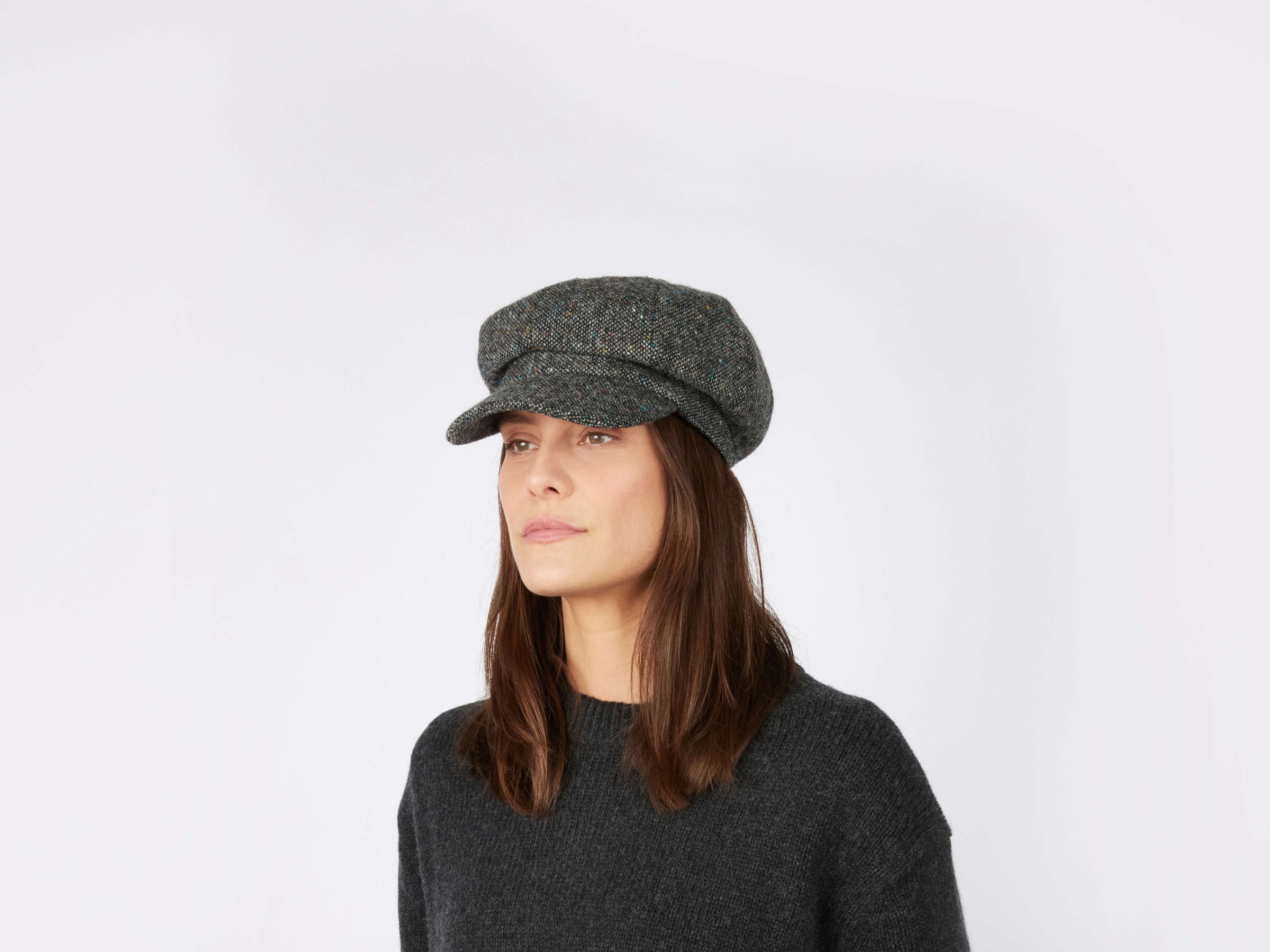 A woman with straight brown hair wears a Hanna Hats Glenveagh Cap Tweed, featuring an eight-panel design, along with a dark sweater. She stands against a light background, looking slightly to the side with a neutral expression.