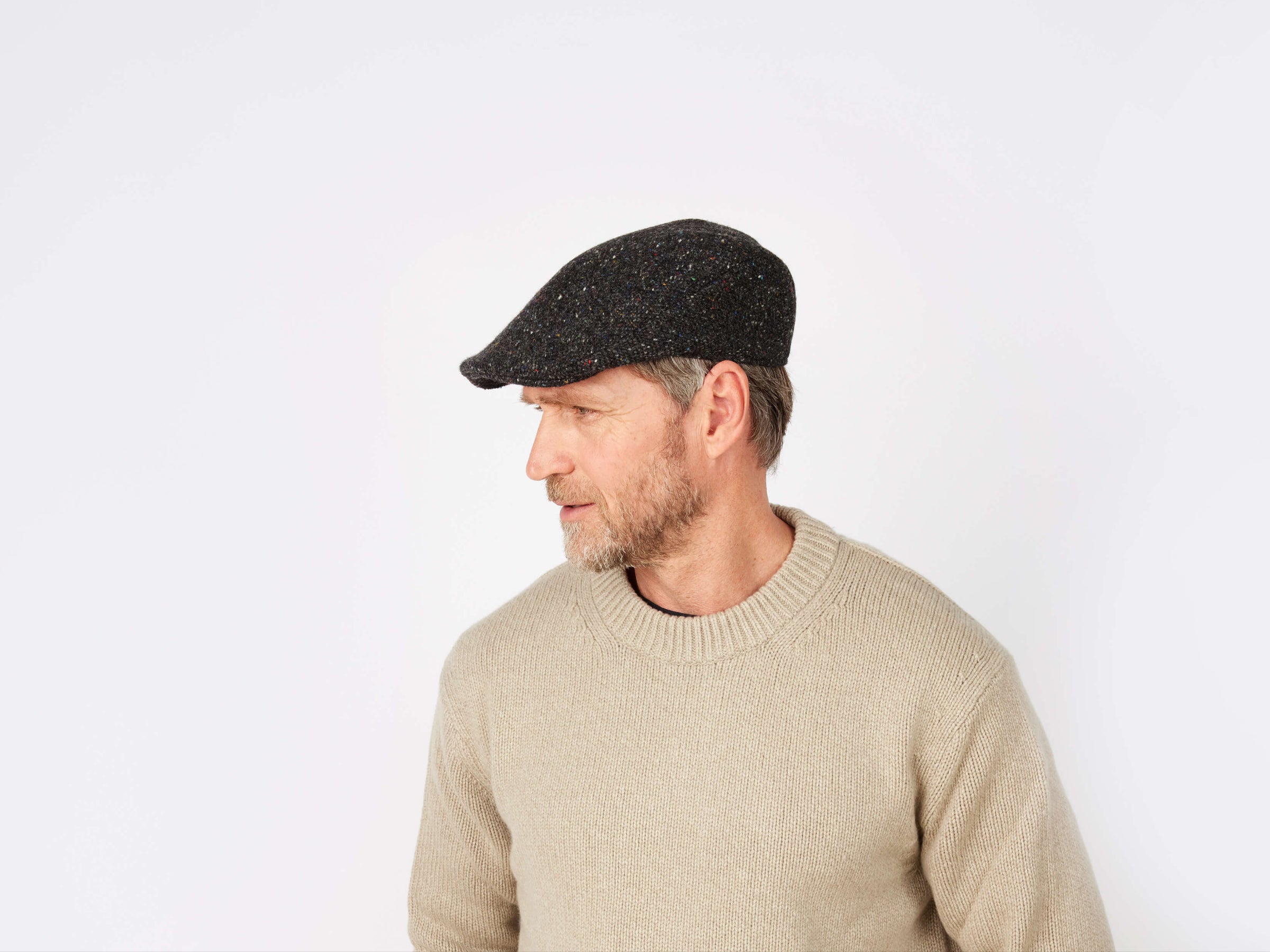 A man in a beige sweater and a tailored fit Hanna Hats Donegal Touring Cap Tweed looks to his left against a plain white background.