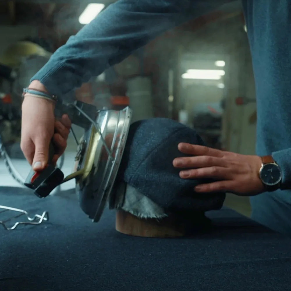 A person uses a steam iron to shape a dark fabric hat on a wooden mold, with one hand guiding the fabric and the other holding the iron. The background shows a workshop setting.
