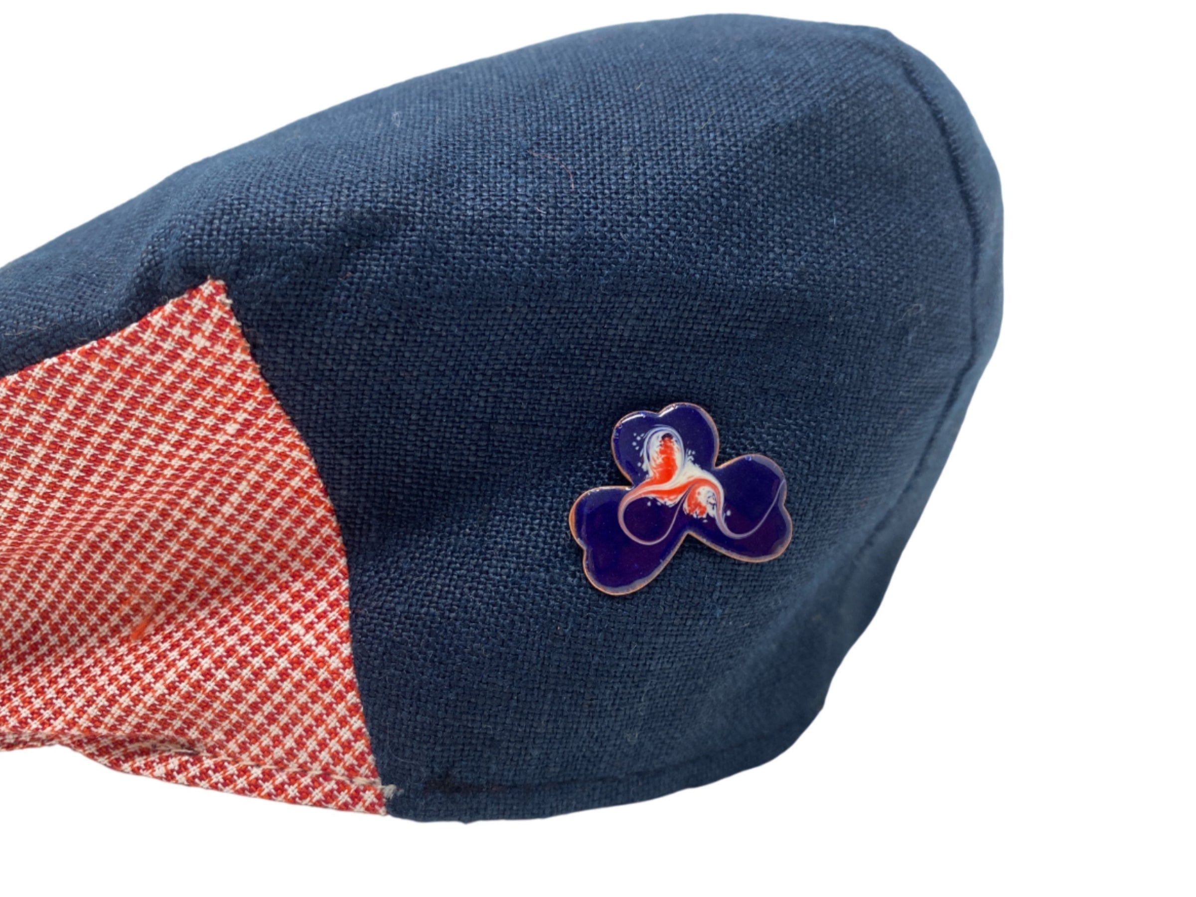 A Hanna Hats of Donegal Ltd navy blue and red checkered cap features a Meab Enamels Lucky Shamrock Pendant Bundle enamel pin on the front, highlighting unique handmade jewellery design, displayed against a white background.