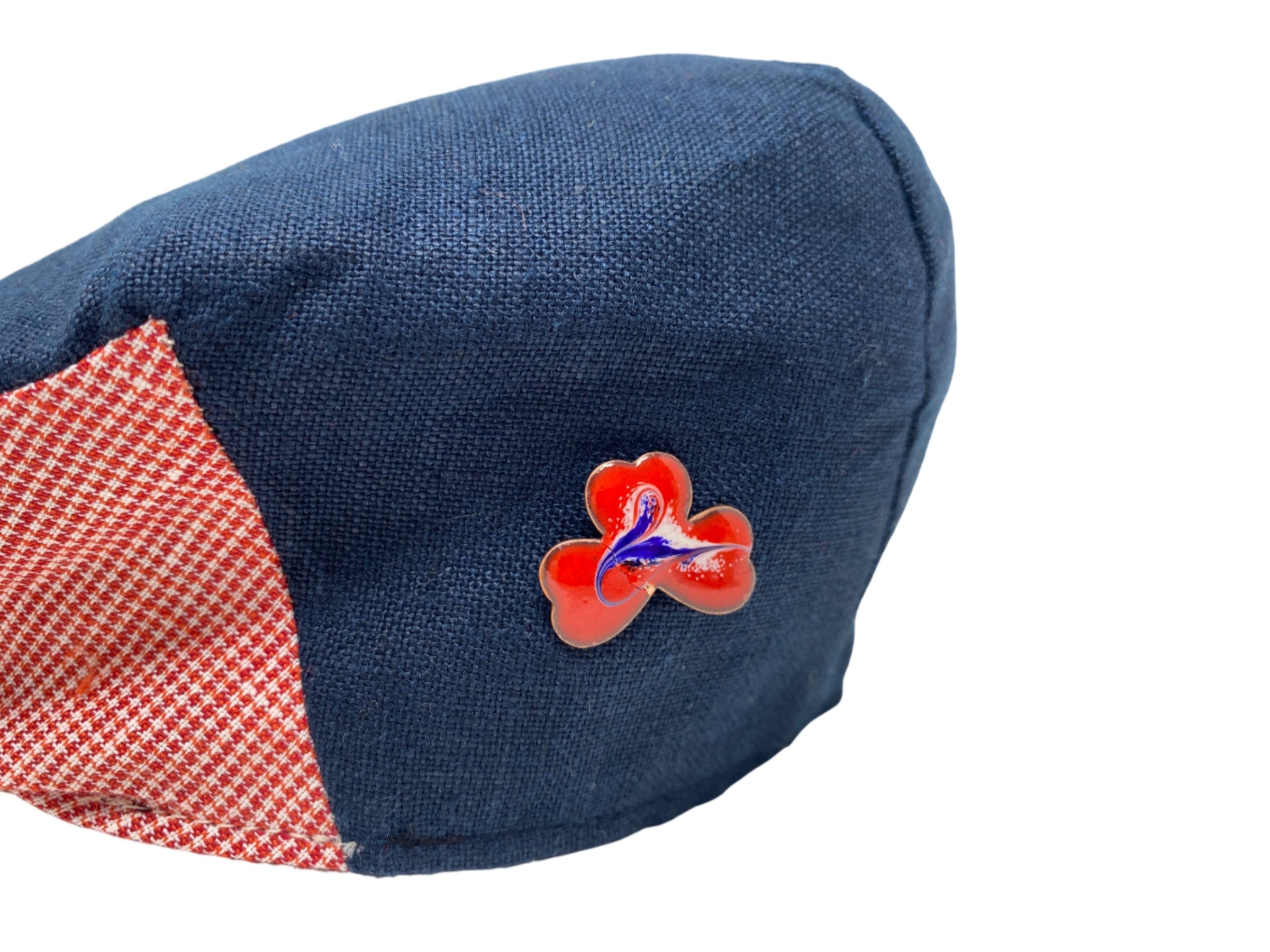 A close-up of a blue fabric cap by Hanna Hats of Donegal Ltd, featuring a red and white checkered section and a red clover pin inspired by the Meab Enamels Lucky Shamrock Pendant Bundle, set against a white background.