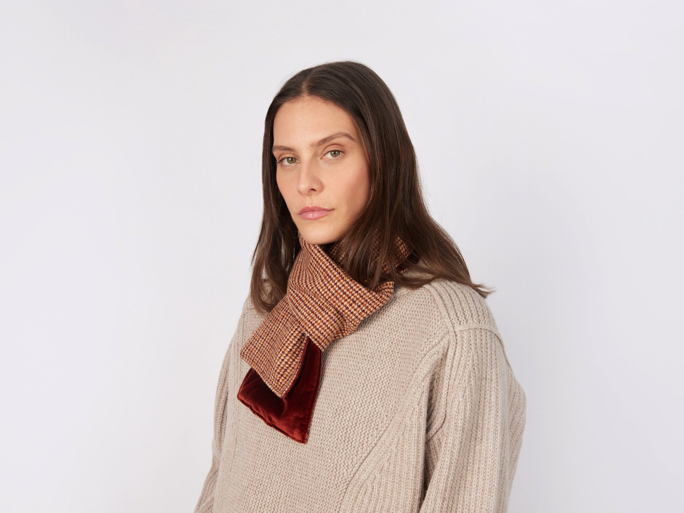 A woman with long brown hair models the Hanna Hats Scarf Tweed / Velvet in red and beige checks over a beige knit sweater, standing against a white background and gazing calmly at the camera.