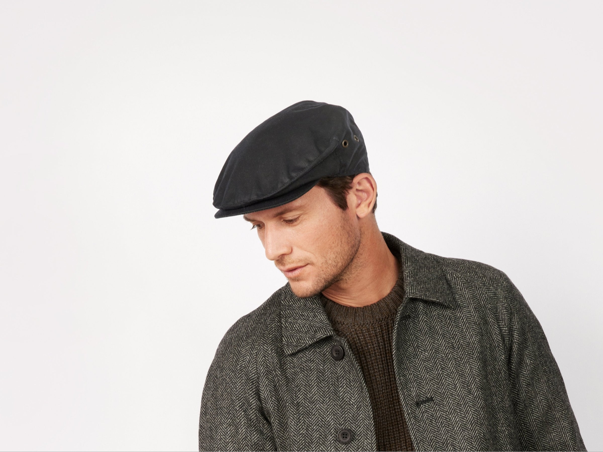 A man wears the Hanna Hats Vintage Cap Wax, paired with a grey textured coat and brown sweater, looking downward against a plain white background.