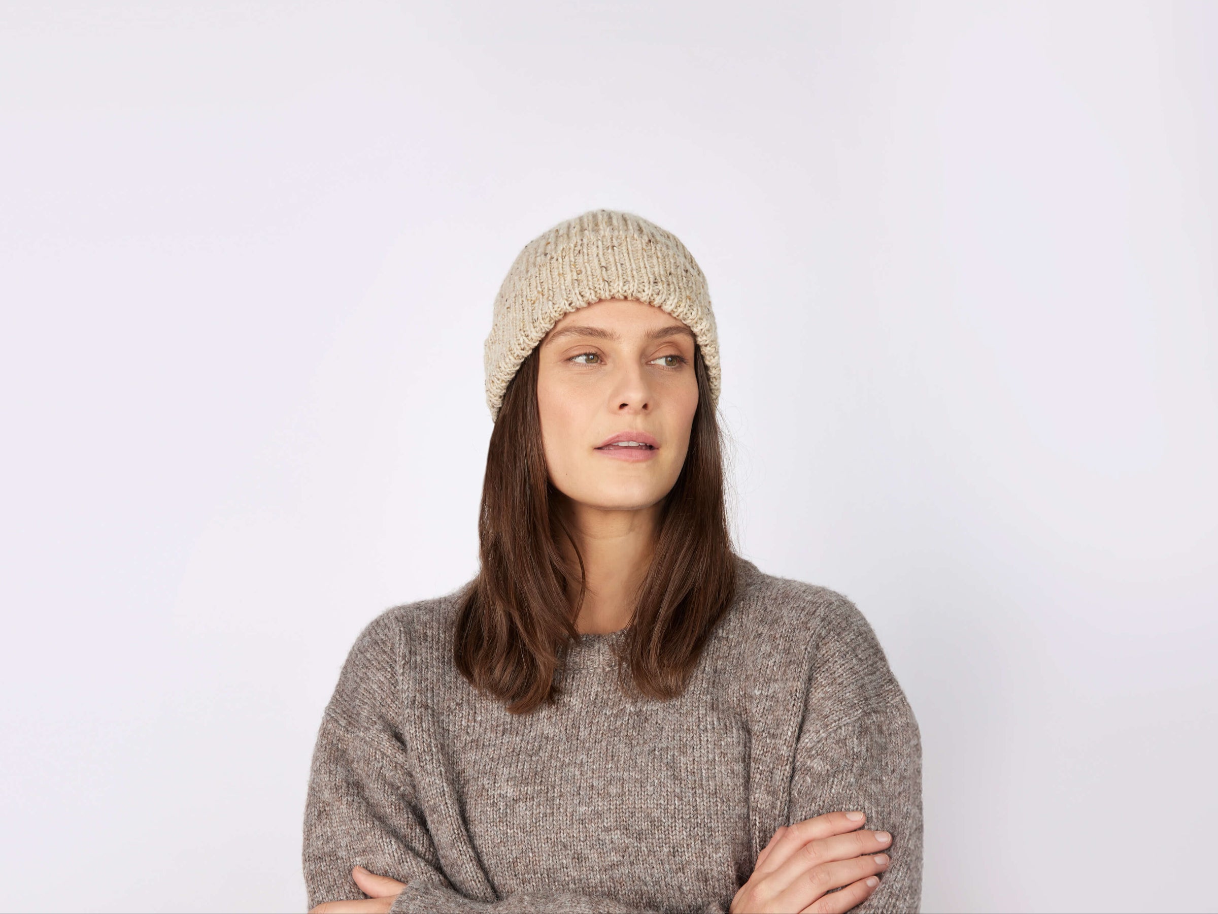 A woman with long brown hair, wearing a Hanna Hats of Donegal Ltd Donegal Beanie and a gray sweater, stands with arms crossed and gazes slightly to the side against a plain light background.